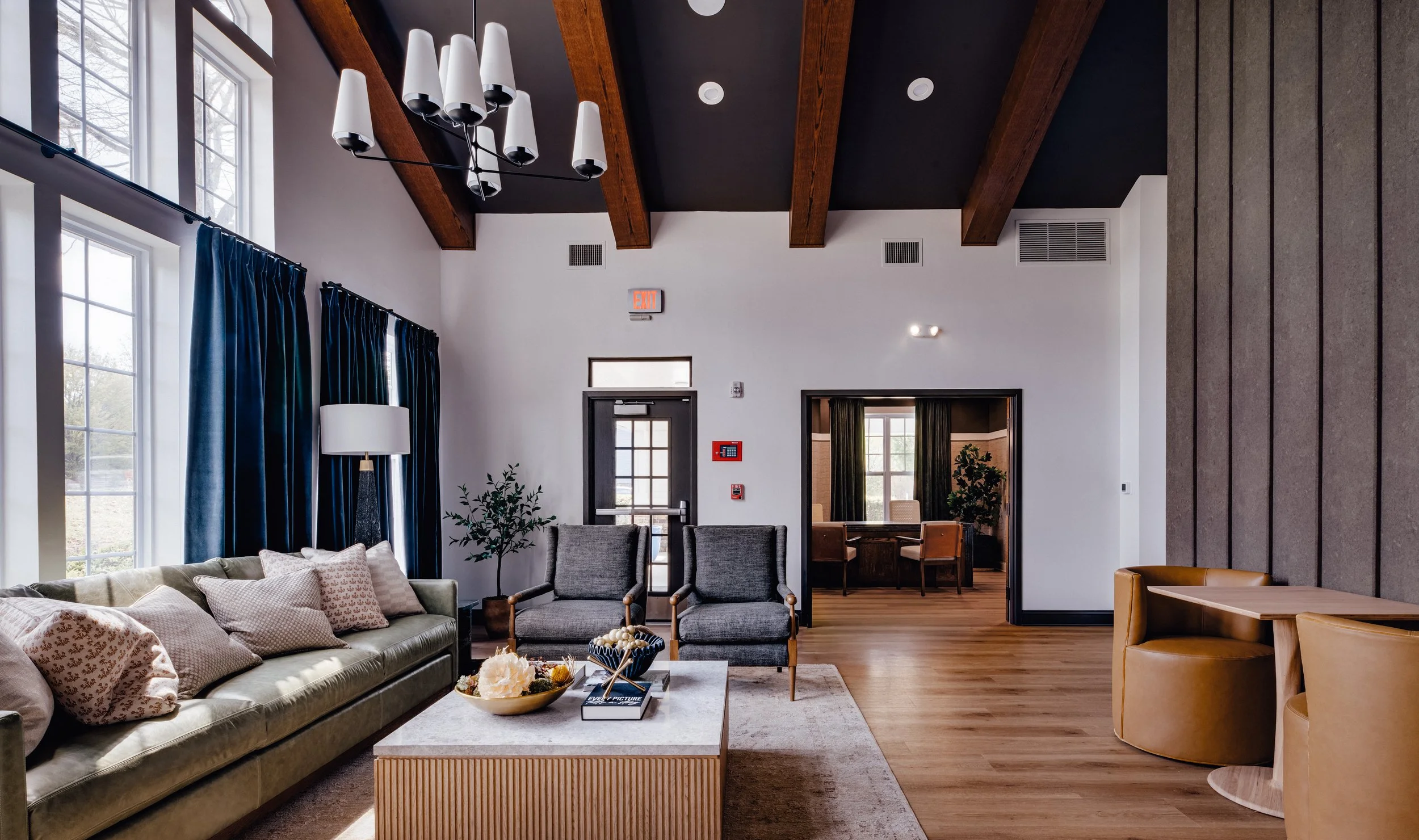 A spacious, modern living room with large windows, dark wood ceiling beams, and a variety of seating including a green sofa with throw pillows, gray armchairs, and a tan armchair. Decor includes a white floor lamp, potted plants, and a coffee table w