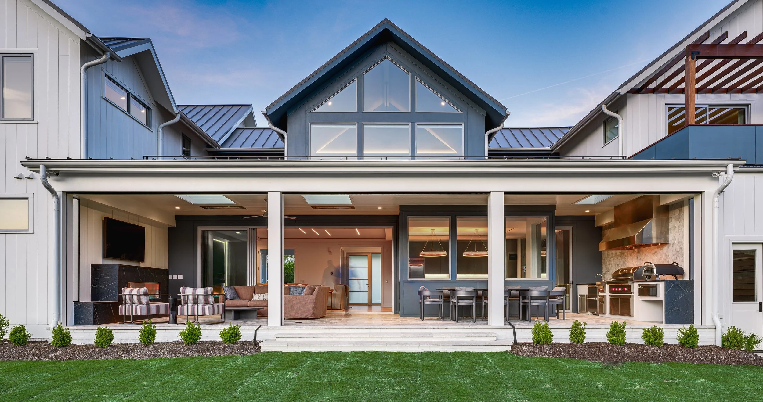Modern house with a large covered patio, outdoor living area, and outdoor kitchen, surrounded by grass and small shrubs.
