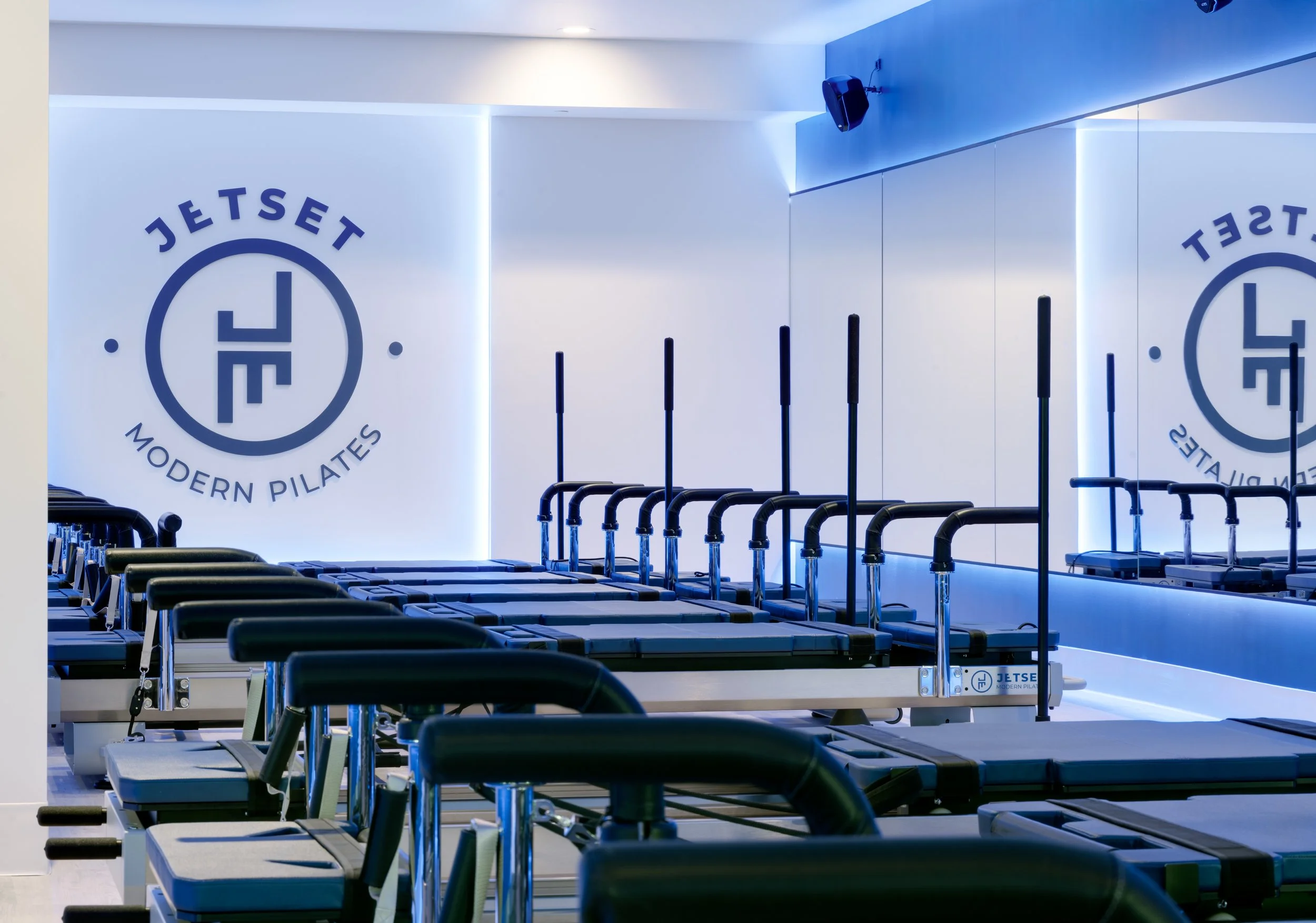An interior view of a modern Pilates studio with rows of reformer machines, mirrors, and a logo on the wall that reads 'Jetset Modern Pilates' with a clock design.