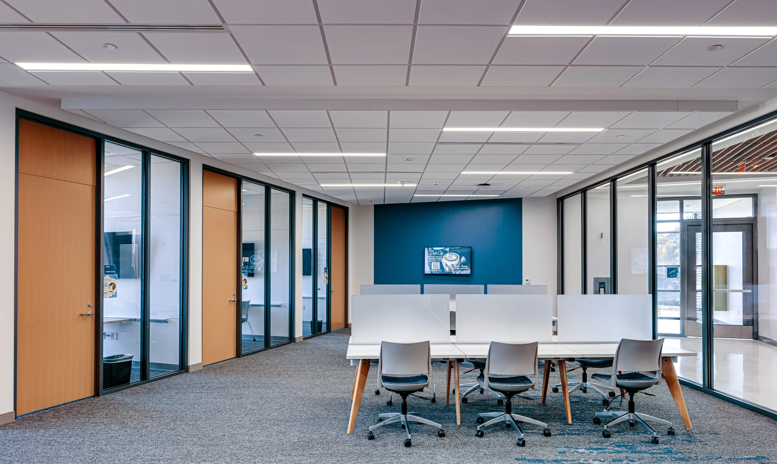 Modern waiting area with white chairs, glass partition walls, a wall-mounted TV, and a ceiling with rectangular panels and long lighting fixtures.
