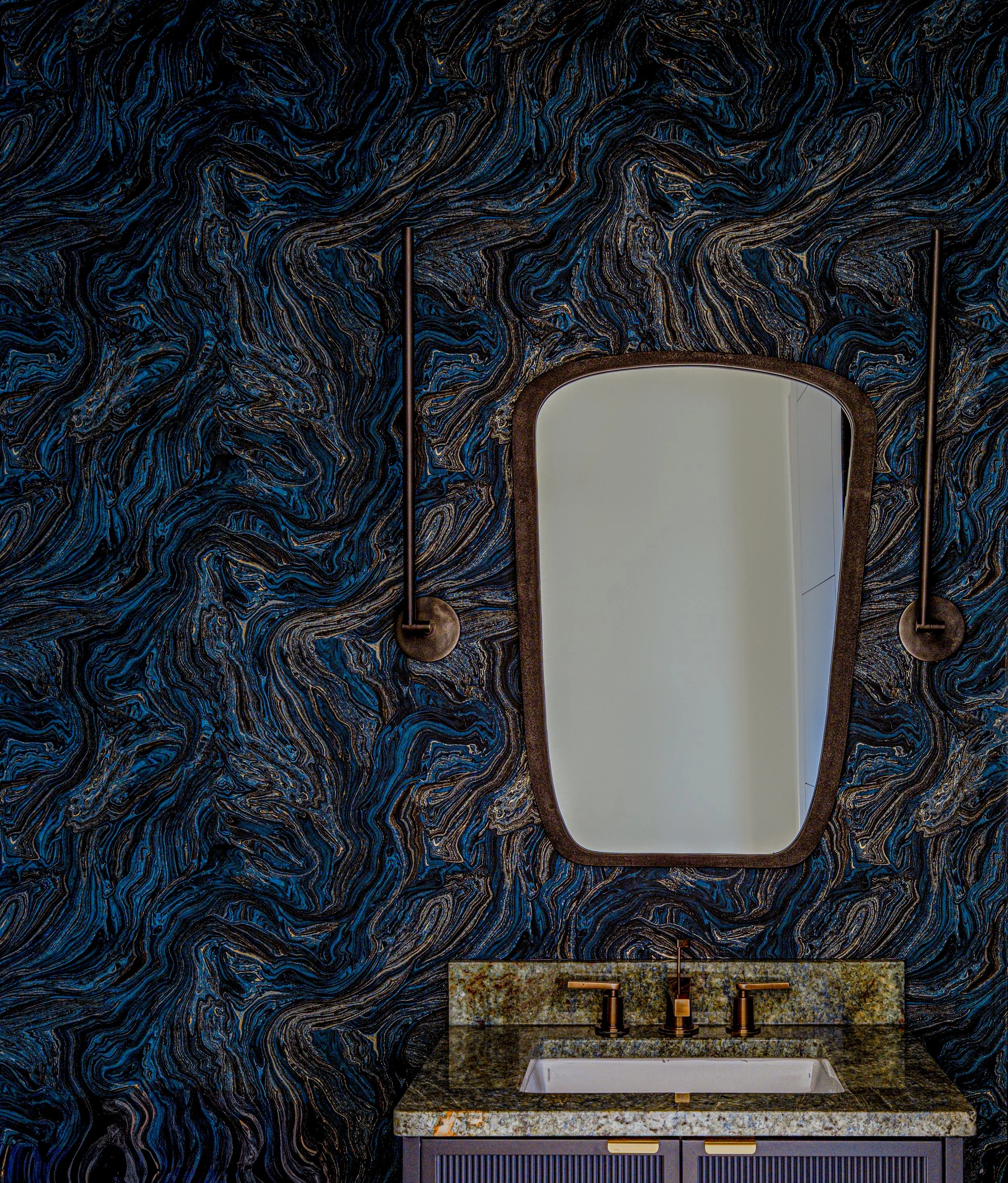 Bathroom with dark marbled wall, rectangular mirror with rounded edges, gold fixtures including a faucet and handles, marble countertop with a blend of tan, cream, and gray, a sink, and vanity drawers in a dark color.
