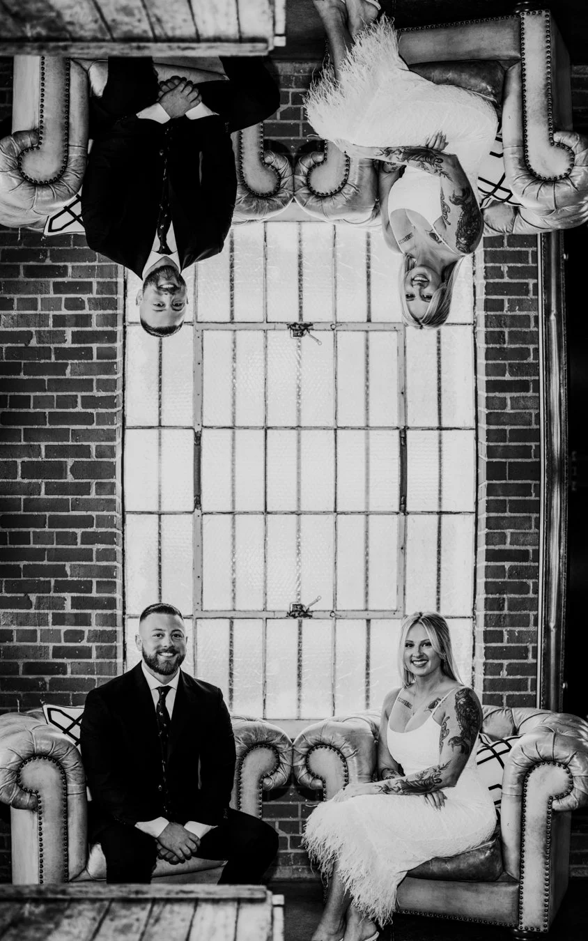 A black and white photo of a man and woman sitting on vintage-style sofas in front of a large window with brick walls on either side. The woman is wearing a white dress with tattoos on her arms, and the man is dressed in a suit.