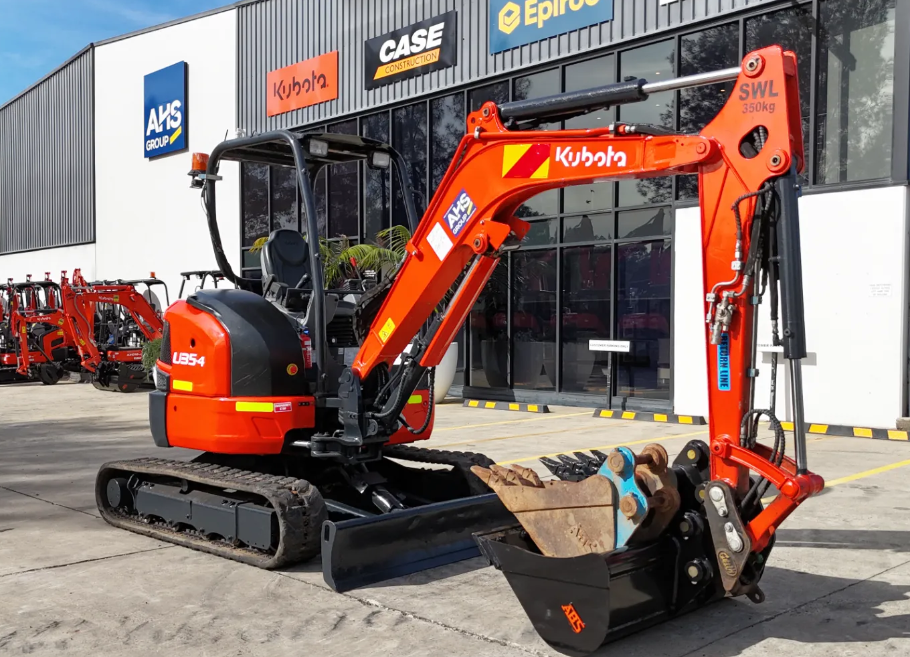 A Kubota compact excavator with a hydraulic arm and bucket, parked outside a building with signs for AYS Group, Kubota, CASE Construction, and Epiroc.