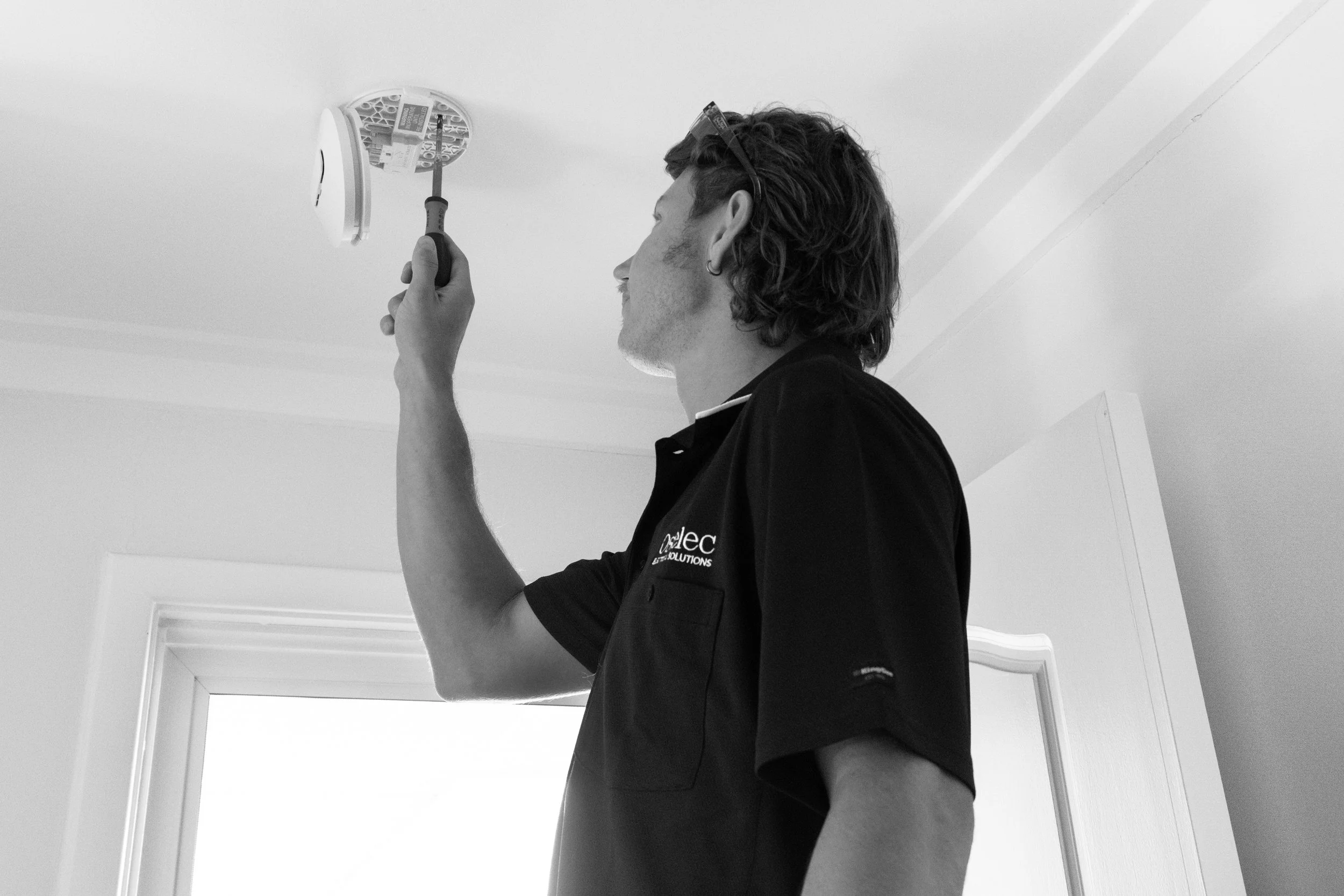 A technician uses a screwdriver to work on a smoke detector on a ceiling in a residential room.