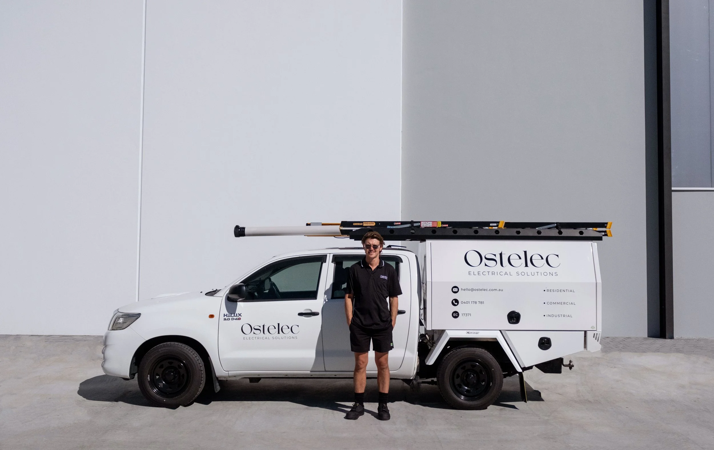 A man standing next to a white service van with the company name 'Ostelec Electrical Solutions'. The man is wearing sunglasses, a black shirt, and shorts. The van has a ladder on top and contact information printed on the side.