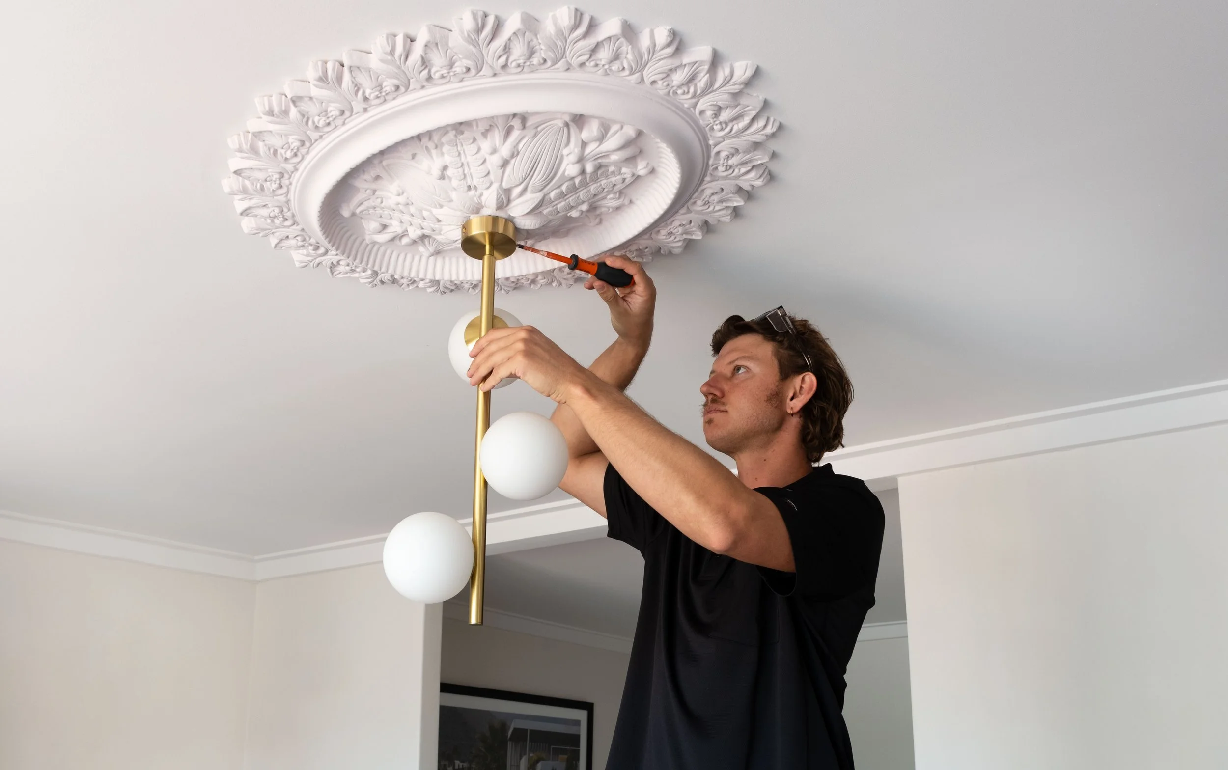 A man installing a decorative ceiling light fixture with ornate molding on the ceiling and white spherical bulbs on a gold rod.