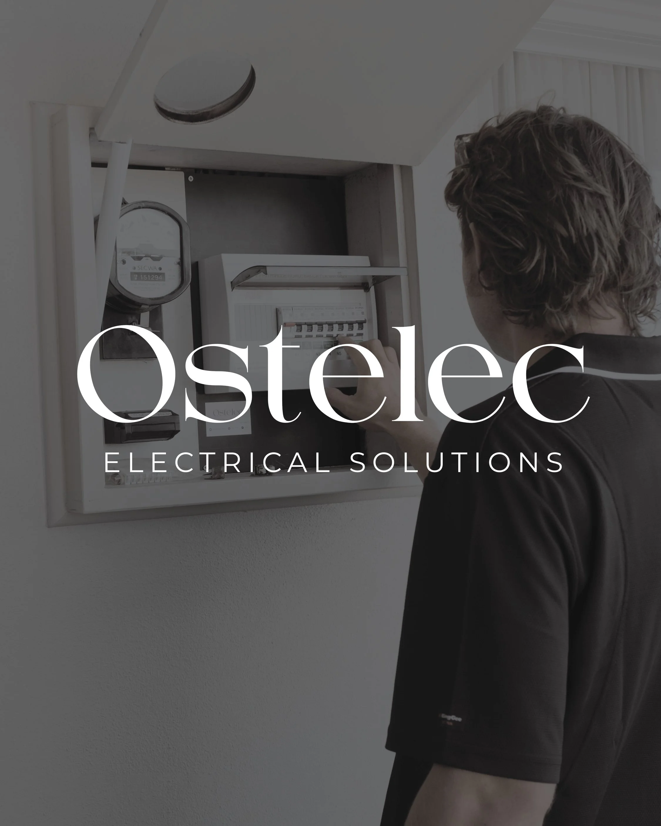 A person with wavy hair wearing a black shirt and light-colored shorts stands in front of an electrical panel or circuit breaker box mounted on a white wall.