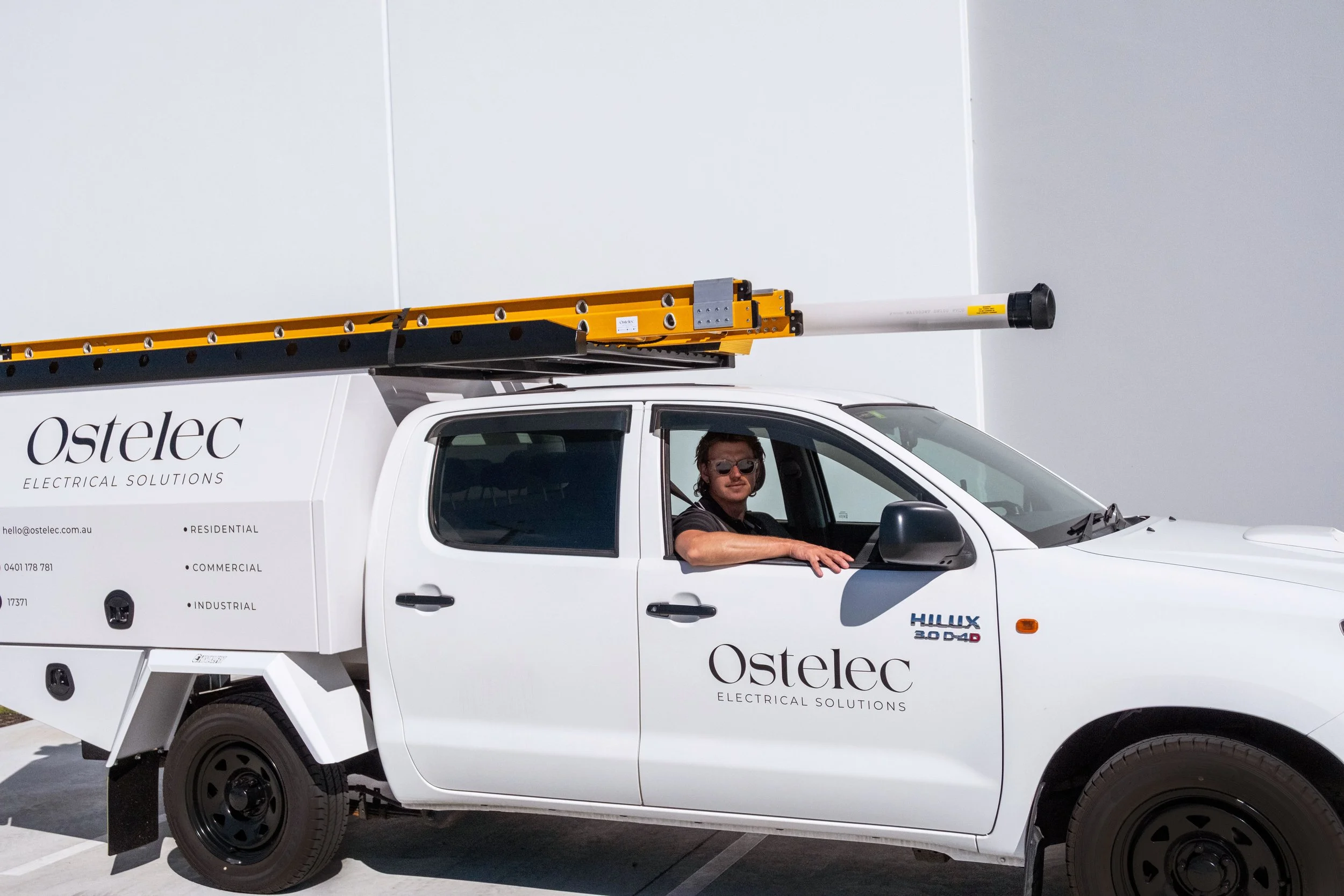 A man sitting in a white pickup truck labeled 'Ostlec Electrical Solutions' with a yellow and black equipment mounted on top. The truck has company details and contact information on the side.