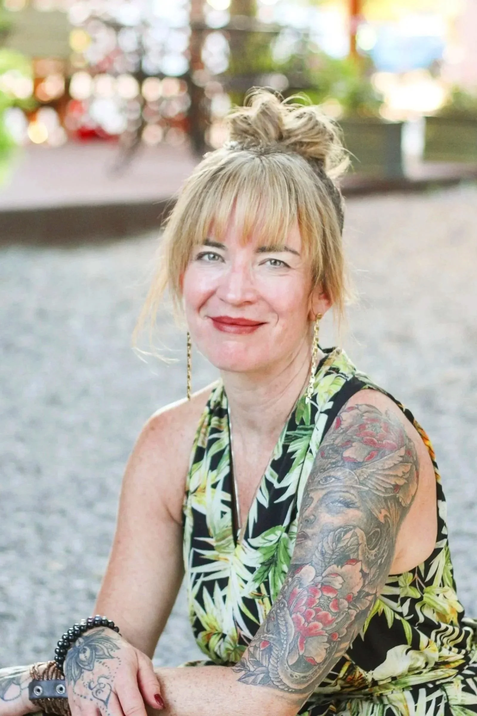 A woman with blonde hair styled in a messy bun, wearing a sleeveless tropical print dress, sitting outdoors on a gravel surface, smiling at the camera, with visible tattoos on her arms and colorful background with blurred trees and lights.