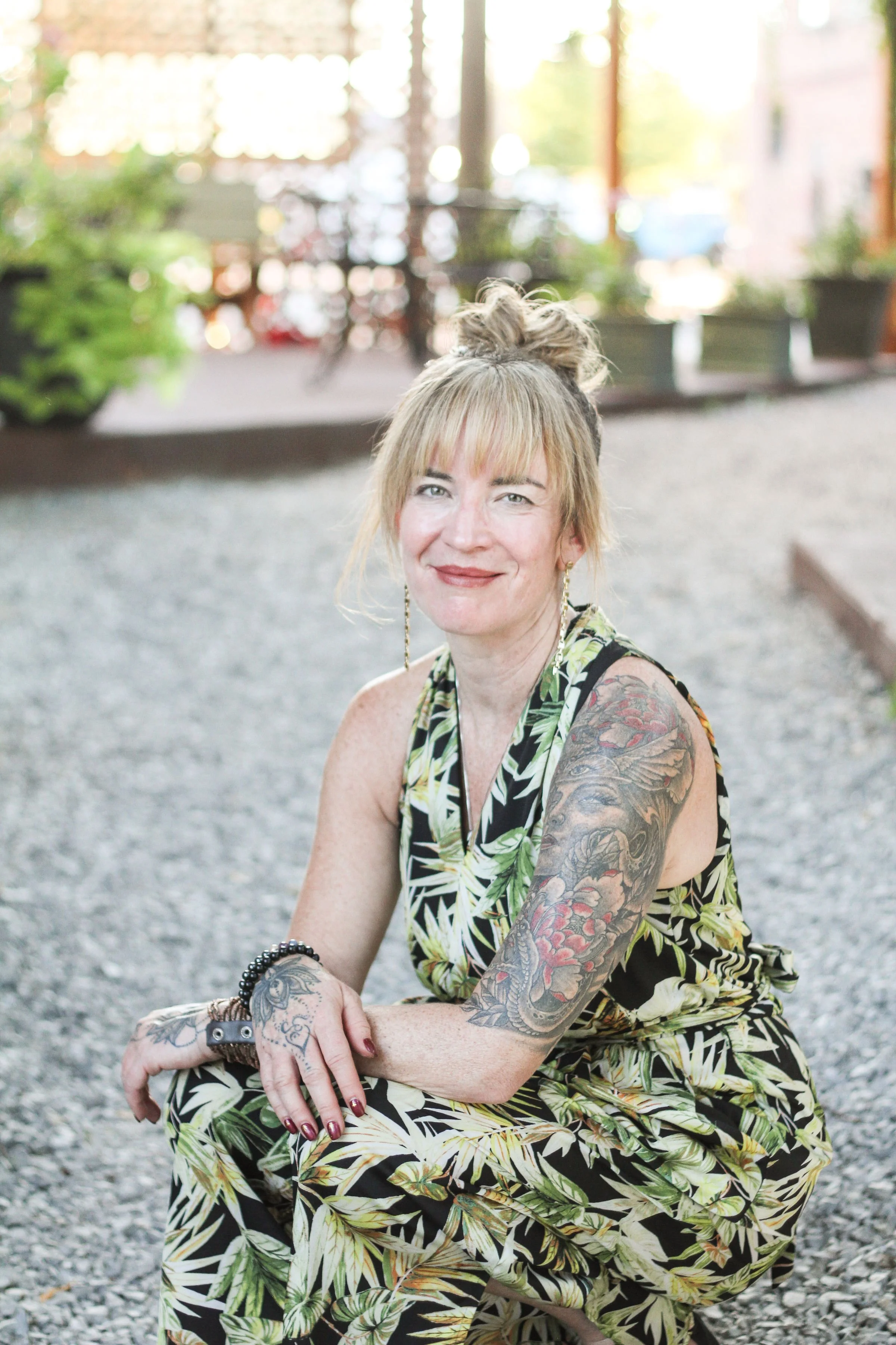 A smiling woman with tattoos on her arms, wearing a matching floral patterned sleeveless dress, sitting outdoors with a blurred background of greenery and sunlight.