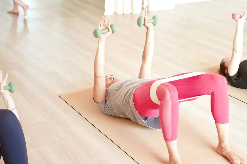 People doing a group exercise or yoga class, lying on mats, lifting small dumbbells in a bright, spacious studio.