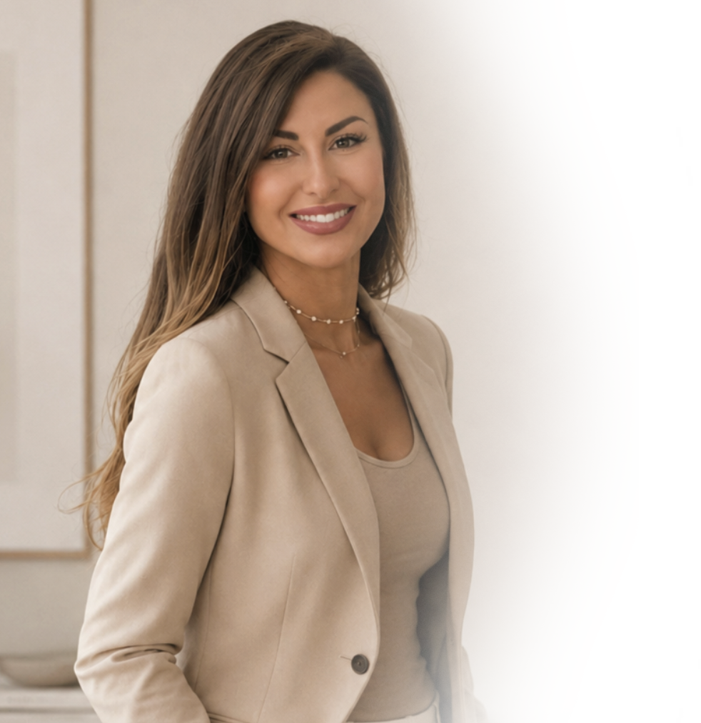 A woman with long brown hair, wearing a beige blazer and a beige top, smiling at the camera in an indoor setting.