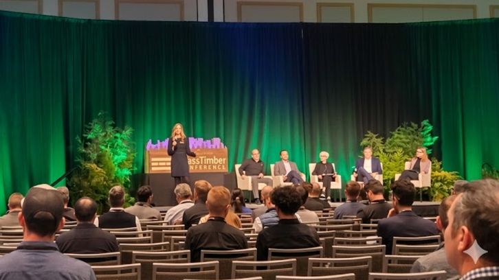 A panel of five people sitting on stage during a conference with a woman speaking at a podium. The background features green and black curtains and trees, with a sign reading "Timber Conference." Audience members are seated facing the stage.