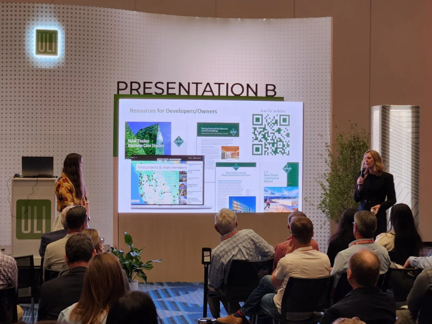 A presentation room with a large screen displaying slides titled 'Resources for Developers/Owners' and images of project case studies and QR codes. A woman in a black outfit holds a microphone and speaks to an audience seated in front of her, with another woman standing to the left near a podium marked 'ULE'.
