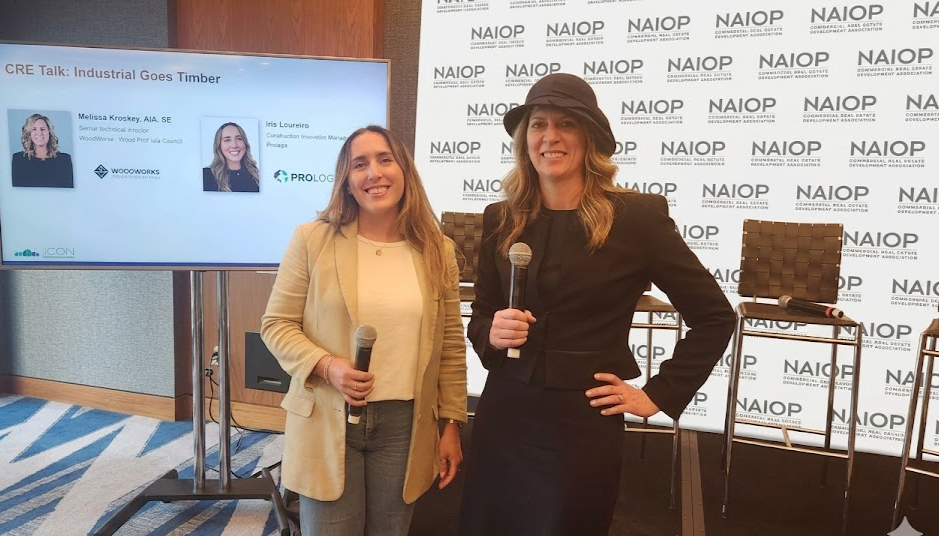 Two women holding microphones at a panel discussion with a NAIOP backdrop and a presentation screen, smiling at the camera.