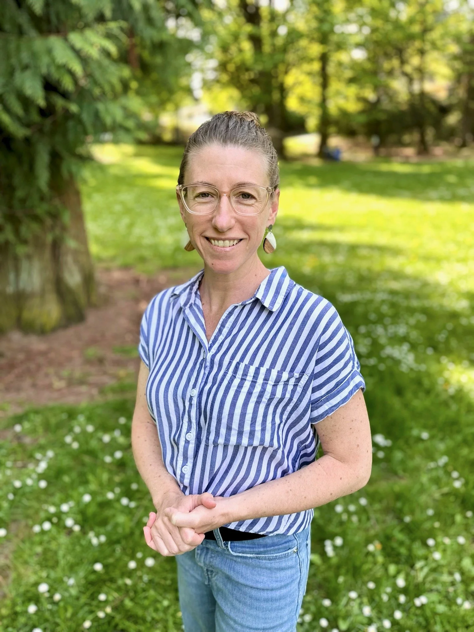 Julie Letchner (a white woman in her 40's) stands in a grassy field, in a 3/4 shot showing thighs to head. She is wearing a blue-and-white striped blouse with jeans, with hair pulled into a messy bun and hands clasped in front of her torso.