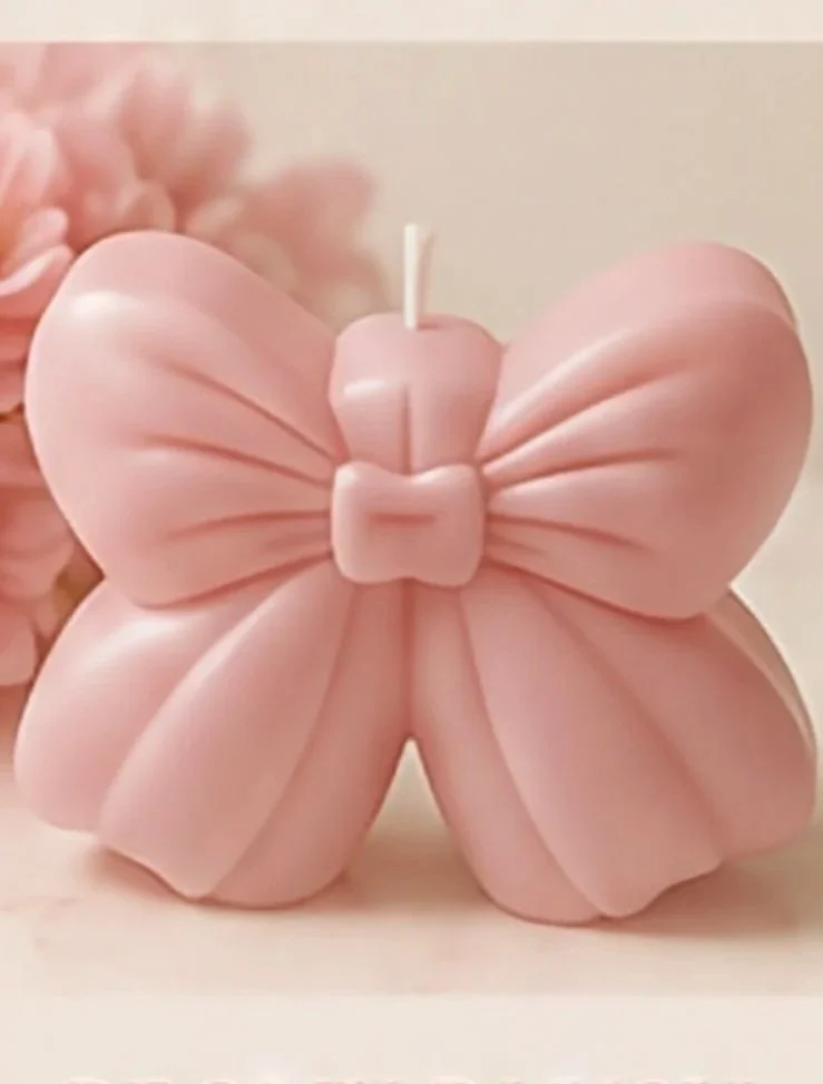 A pink butterfly-shaped candle with detailed wings and a small bow in the center, set on a light surface with pink flowers in the background.