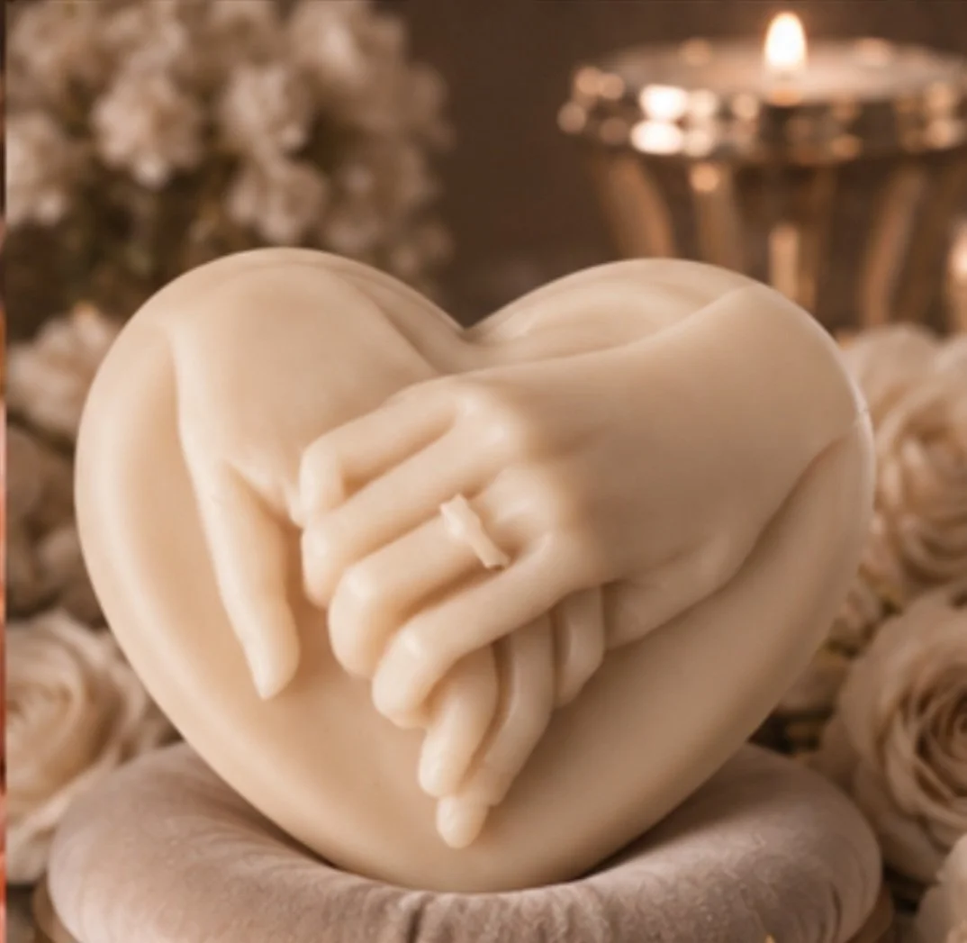A white heart-shaped sculpture with intricately detailed hands holding each other, placed on a rounded base. The background shows blurred white flowers and a lit candle in a decorative holder.