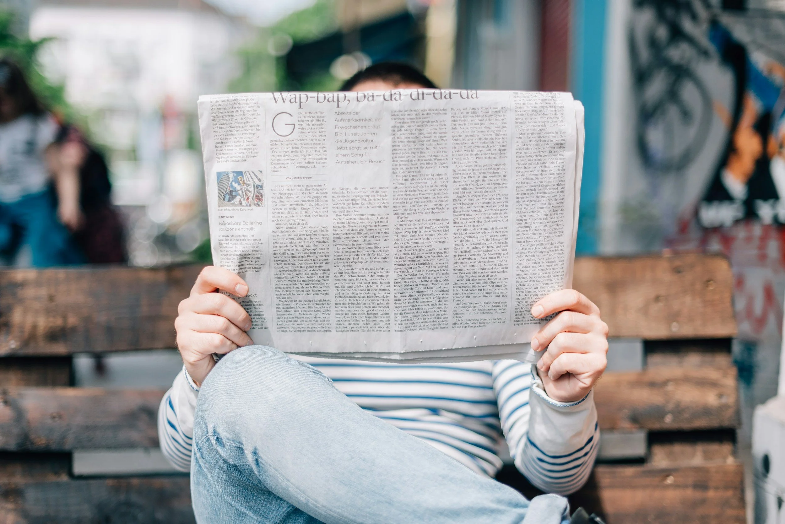 Person sitting outdoors on a wooden bench, reading a newspaper, with their face partially obscured by the paper. They are wearing a striped shirt and light blue jeans, with graffiti-covered walls visible in the background.