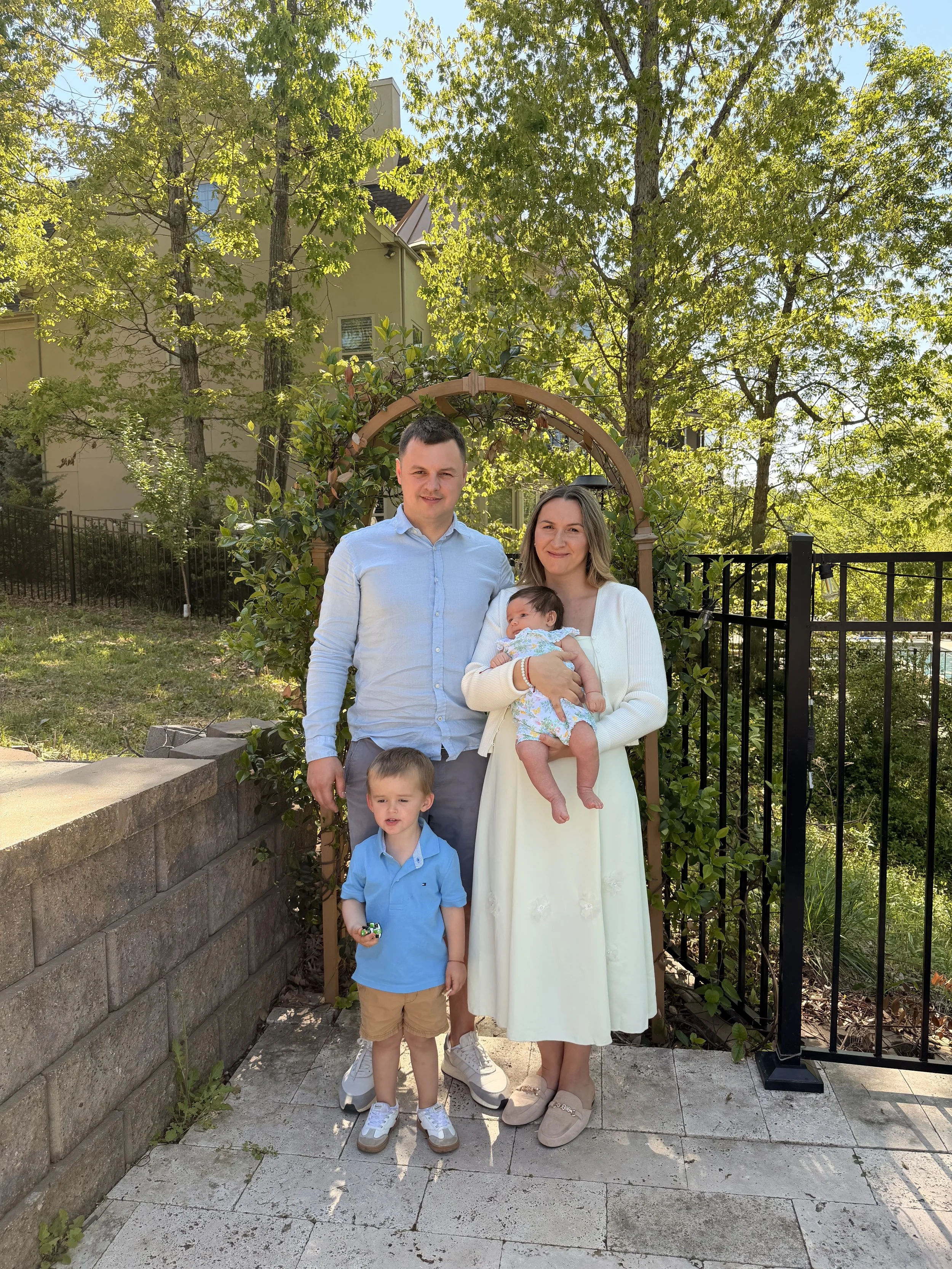 A family of five standing outdoors in front of a garden arch, surrounded by trees. The woman is holding a baby, the man has his arm around her, and two young boys stand in front of them. All are smiling.