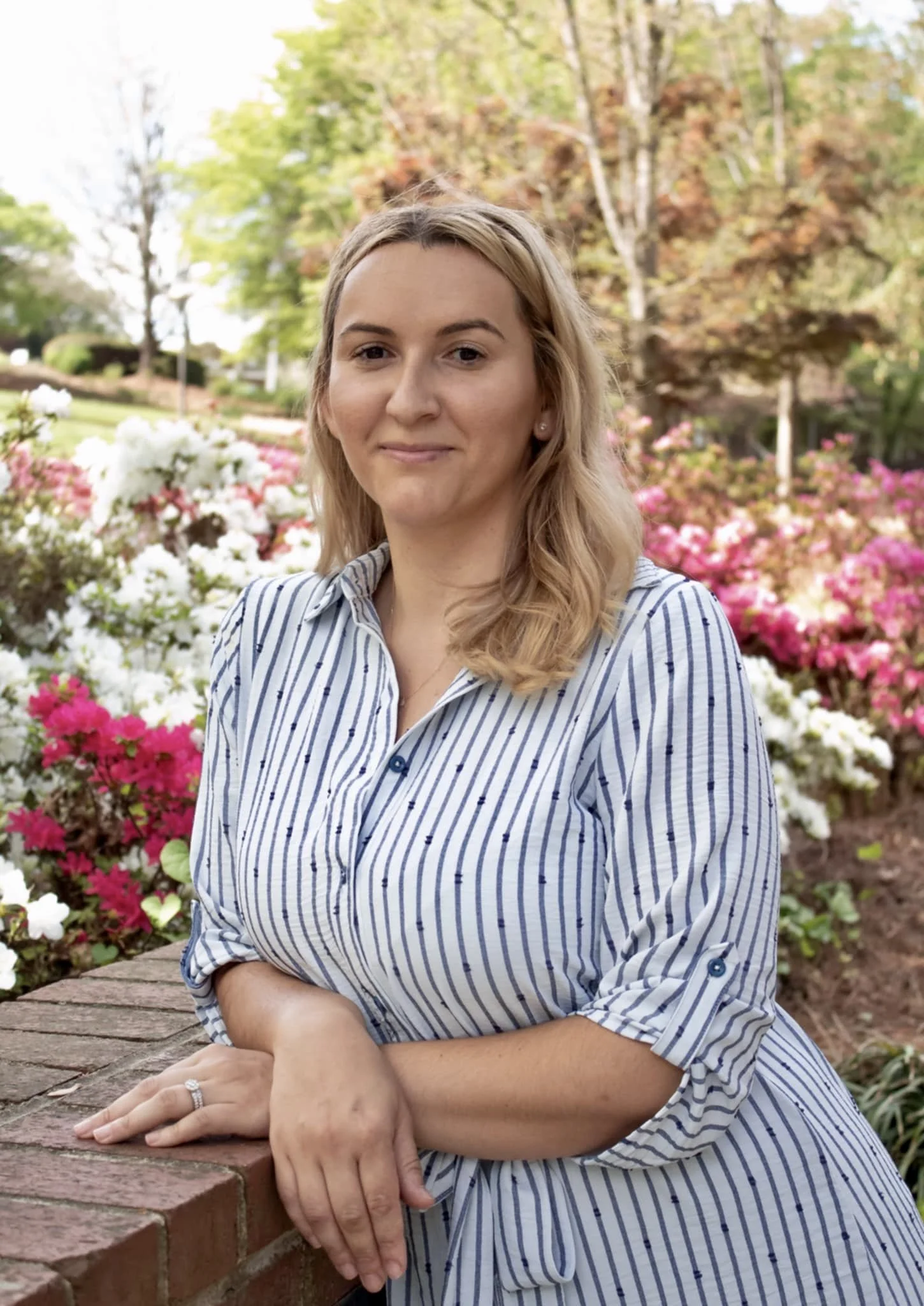 A woman with blonde hair and a striped shirt leaning on a brick ledge outdoors in a garden with pink and white flowers and trees with green and autumn-colored leaves in the background.
