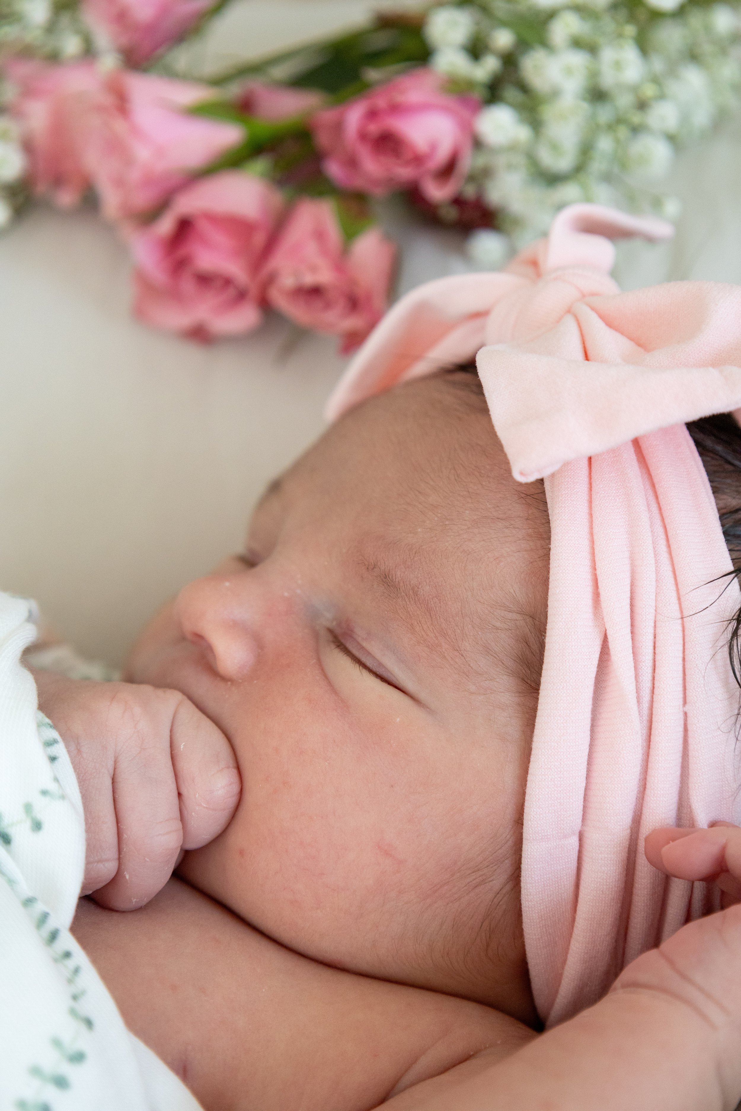 Close-up of a sleeping baby with a pink headband, lying on their side, with a bouquet of pink and white flowers in the background.