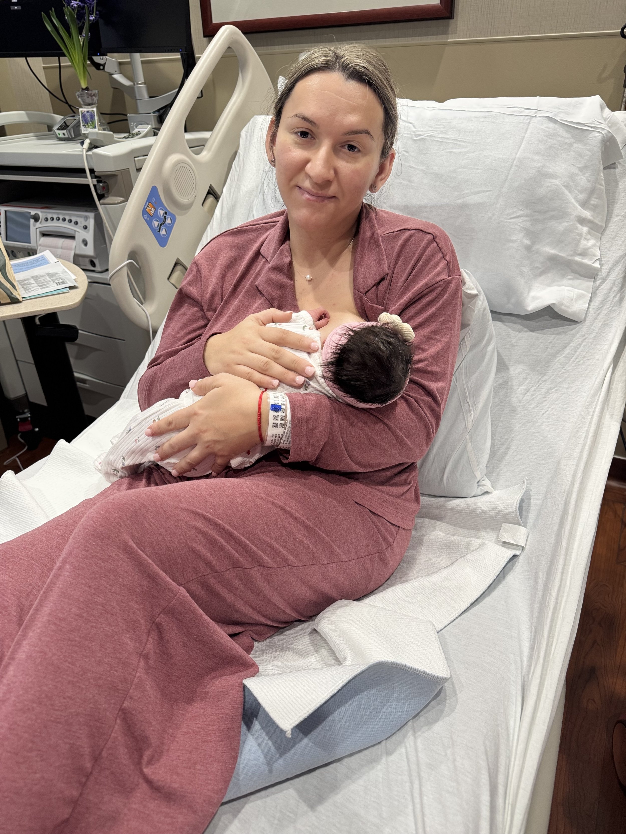 A woman is holding a newborn baby in a hospital bed, smiling at the camera. The woman is wearing a pinkish-brown hospital gown, and the baby is wrapped in a striped blanket. Medical equipment and a monitor are visible in the background.