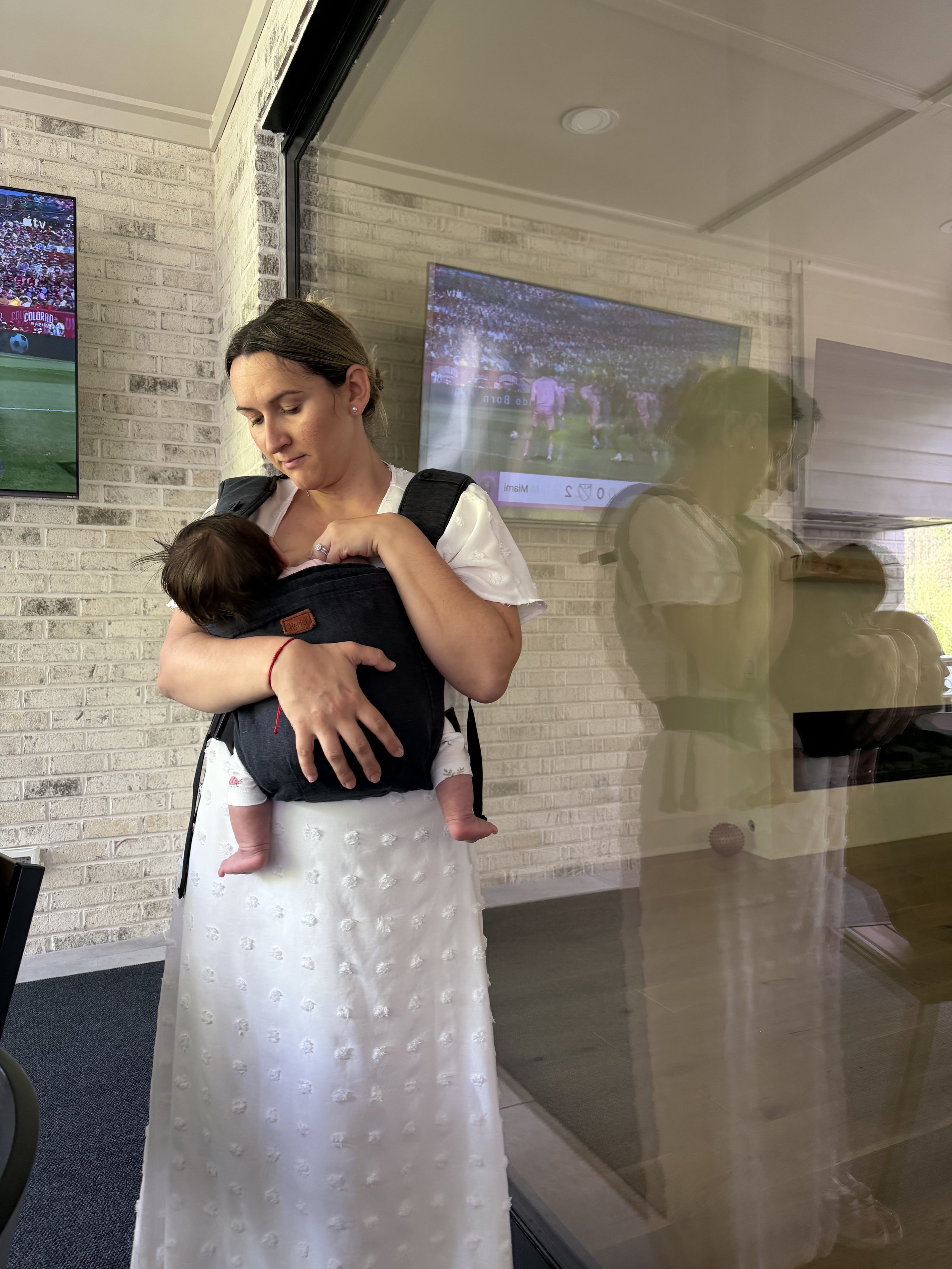 A woman wearing a long white dress is holding a small child in a black baby carrier. The woman is standing indoors in front of a brick wall with two television screens mounted on it.