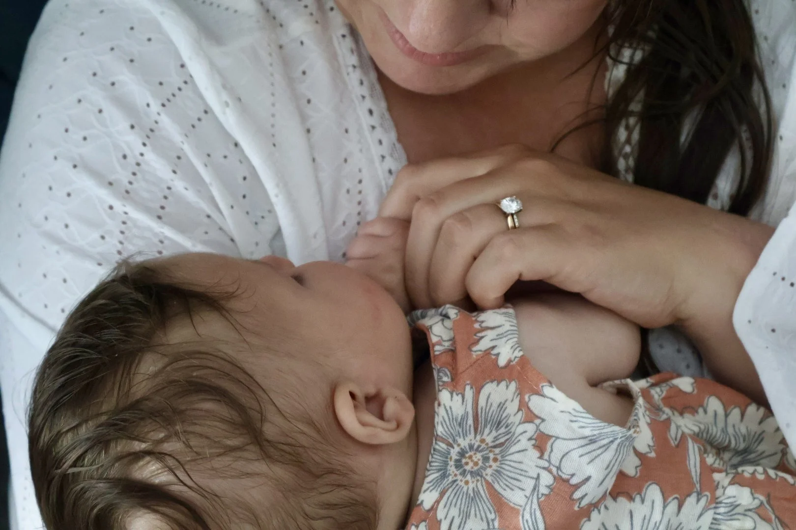 A woman with dark hair wearing a white dress with eyelet details, holding a baby who is breastfeeding. The baby has light brown hair and is wearing a floral-patterned shirt with orange, white, and blue colors. The woman is wearing a large engagement ring with a prominent diamond.