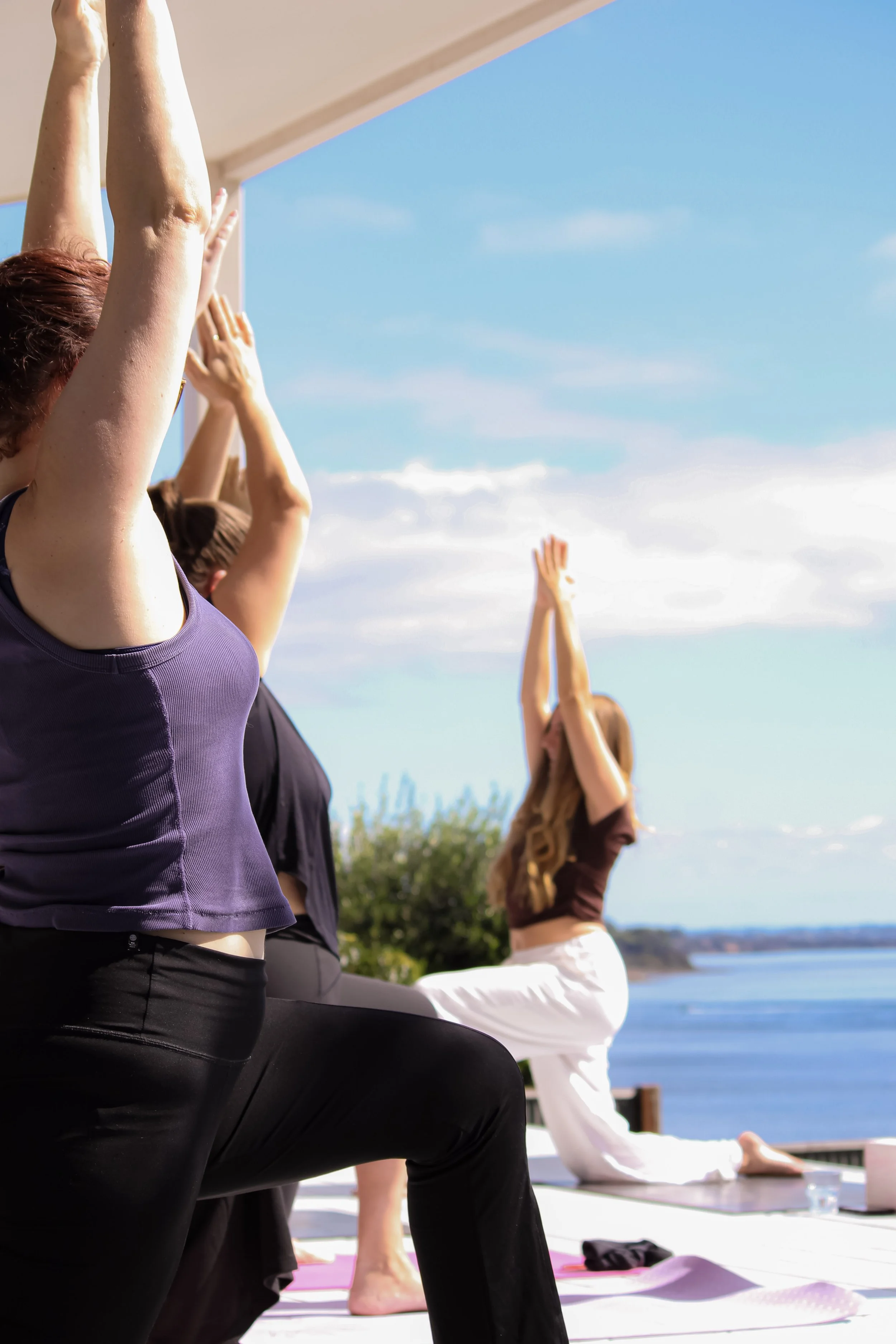 women practicing yoga on the deck of a beach house in San Remo, reaching for the sky with the sea, blue skies and clouds in the background