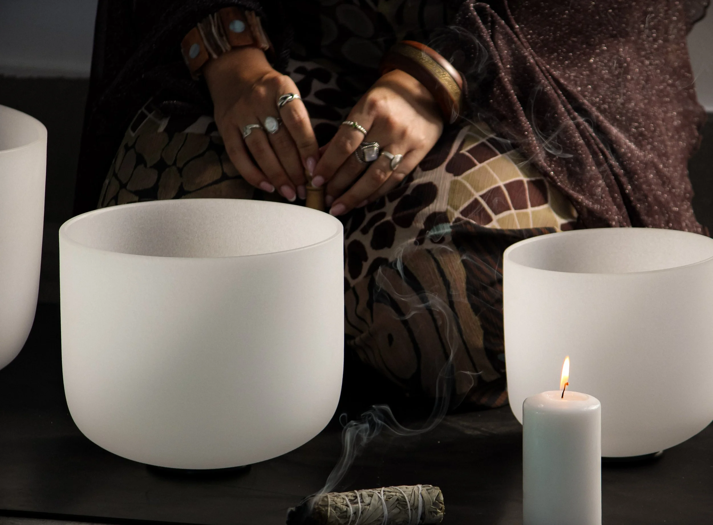 woman's hands with silver rings and bangles, fingers interlocking in front of sound bowls while candles light in a dark moody scene