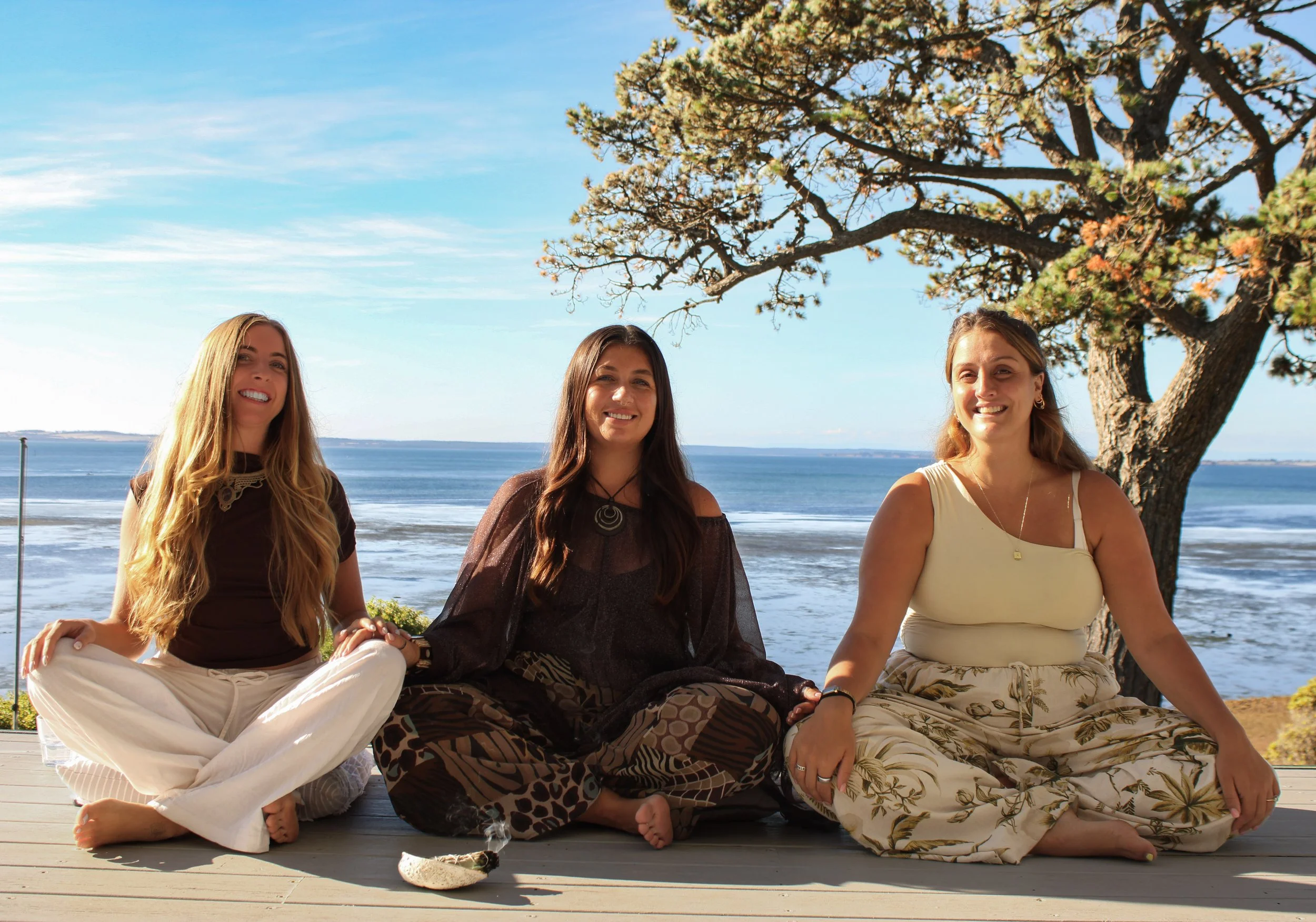 three female business owners hosting a wellness retreat on a deck by the sea, with a tree and blue skies in the background, with smiles and gentle touches