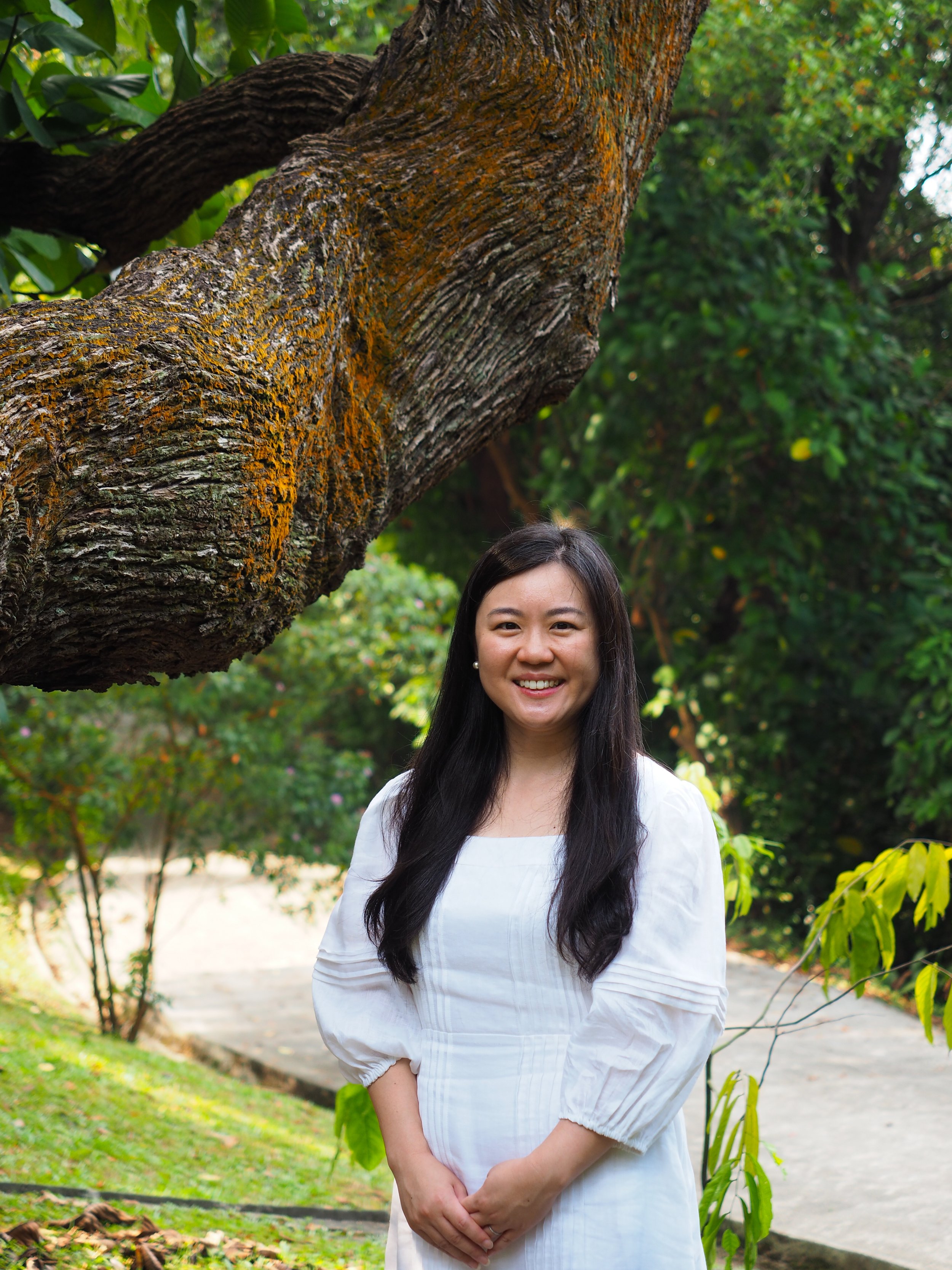 A woman with long dark hair wearing a white dress standing outdoors with green trees and bushes in the background.