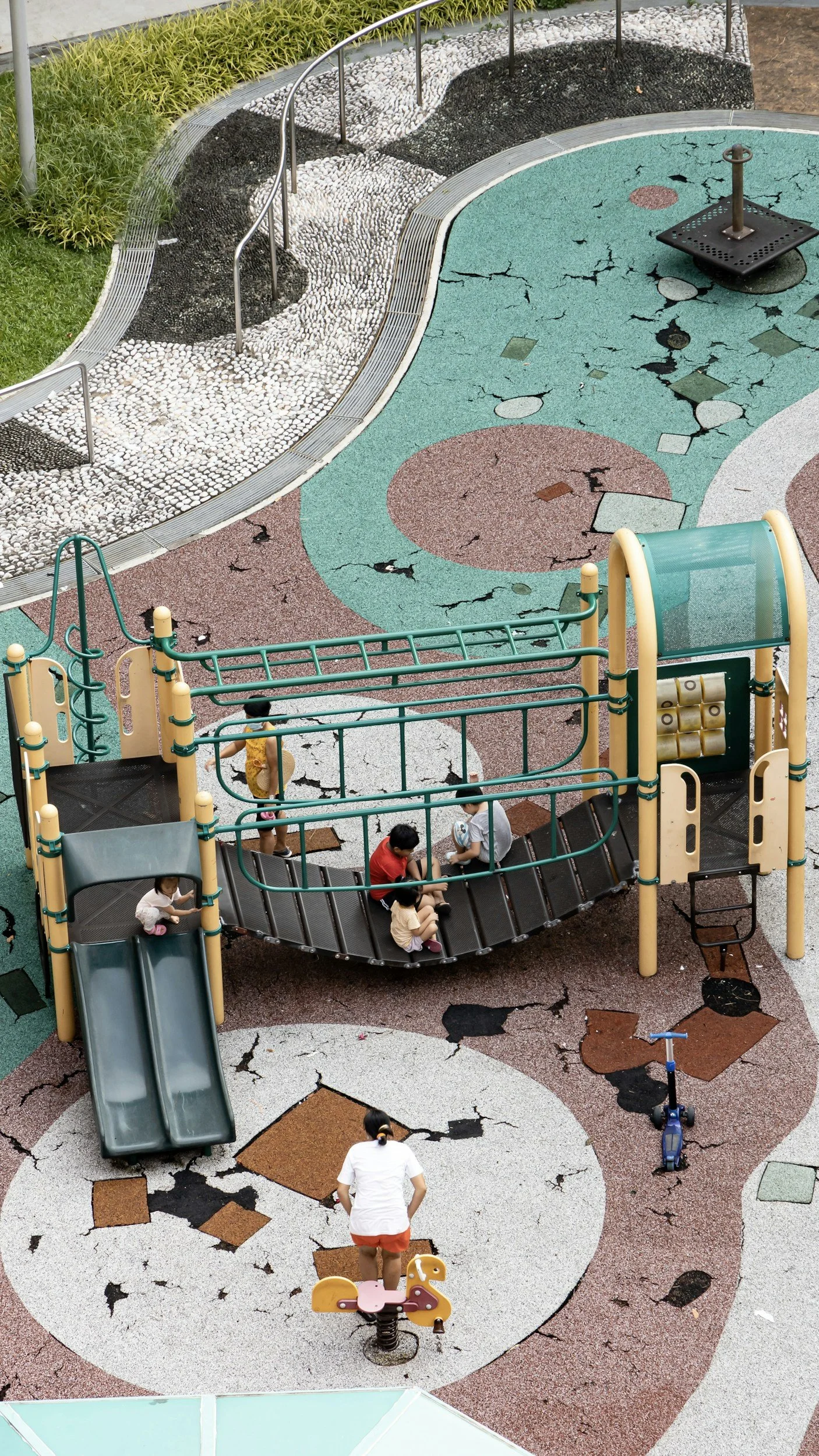 A playground with a cracked rubber surface in colorful designs, a slide, children playing, and surrounding green bushes.