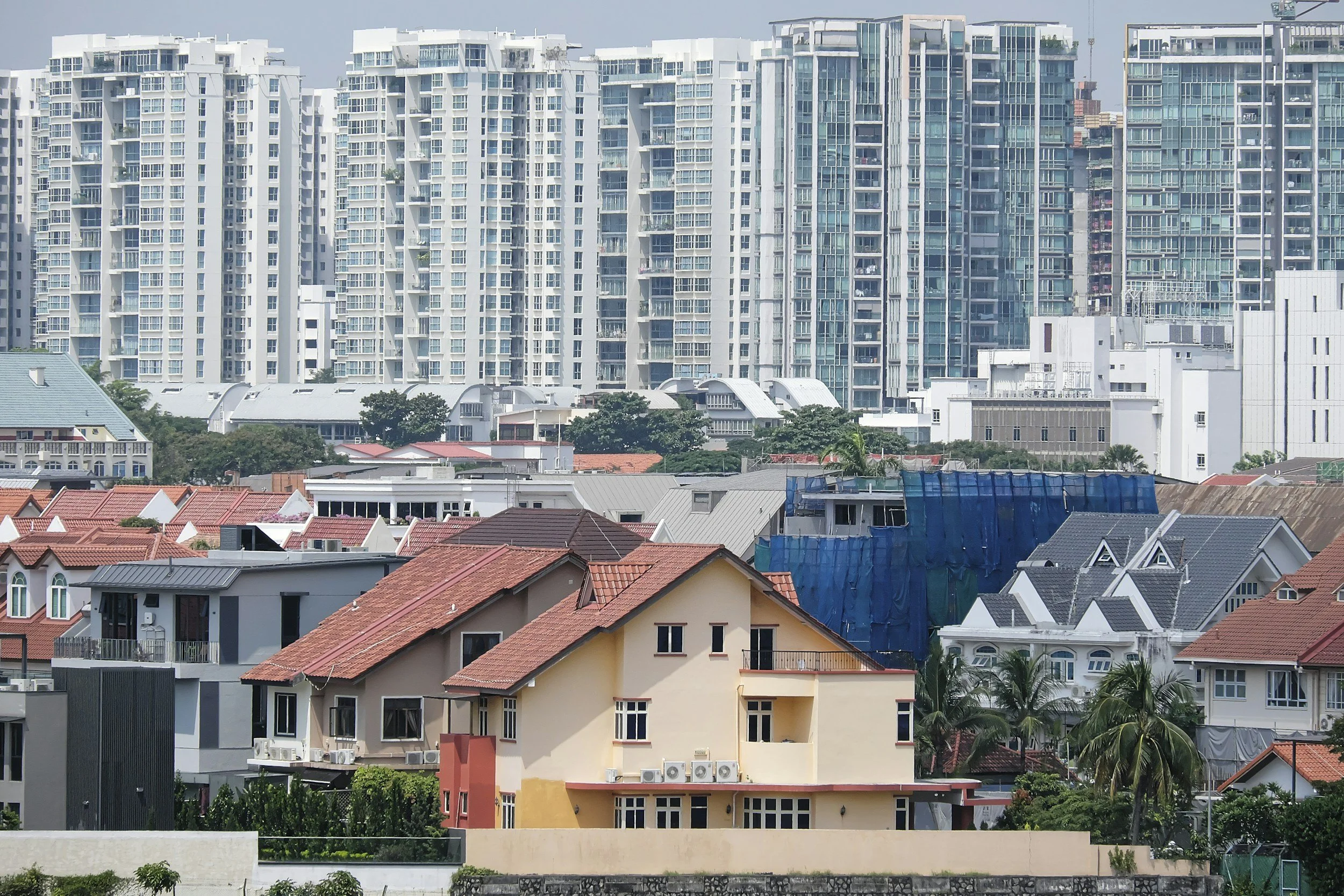 View of a cityscape with a row of large modern high-rise apartments or condominiums in the background and smaller residential houses with red and beige roofs in the foreground. There are some trees and greenery at the bottom.