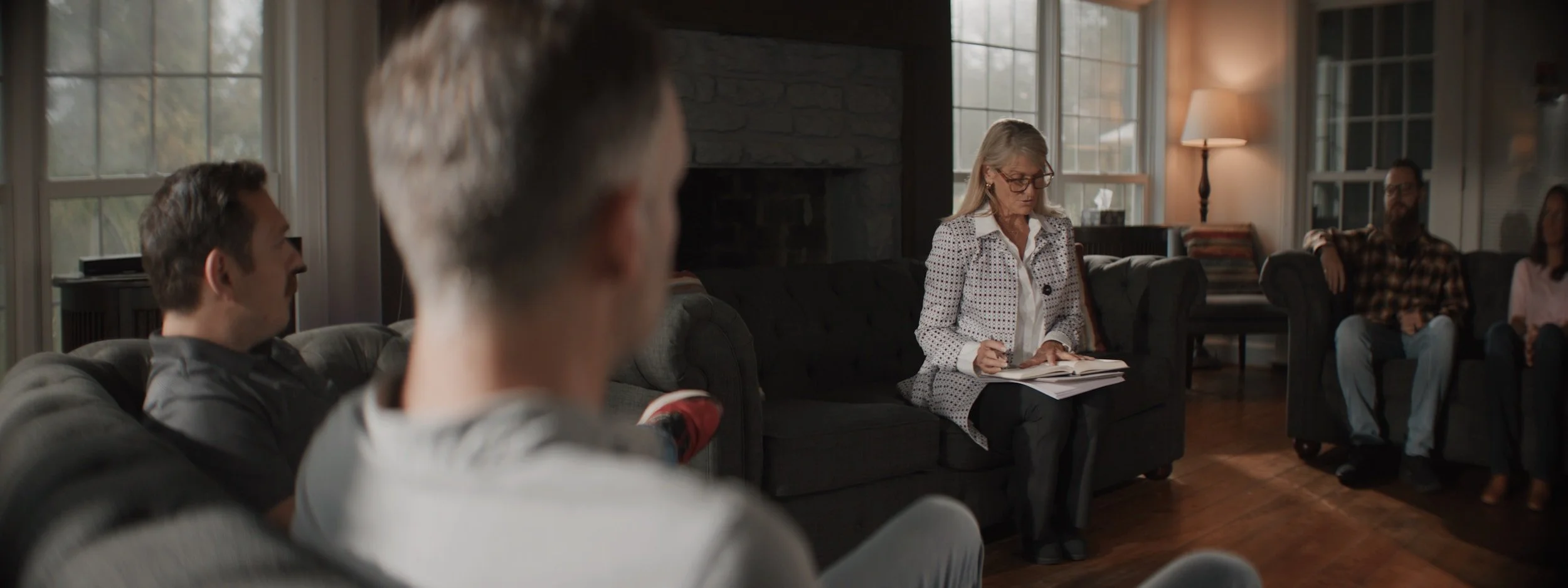 A group therapy session with five adults sitting on couches in a cozy living room, with a woman in a white and black polka dot jacket taking notes.
