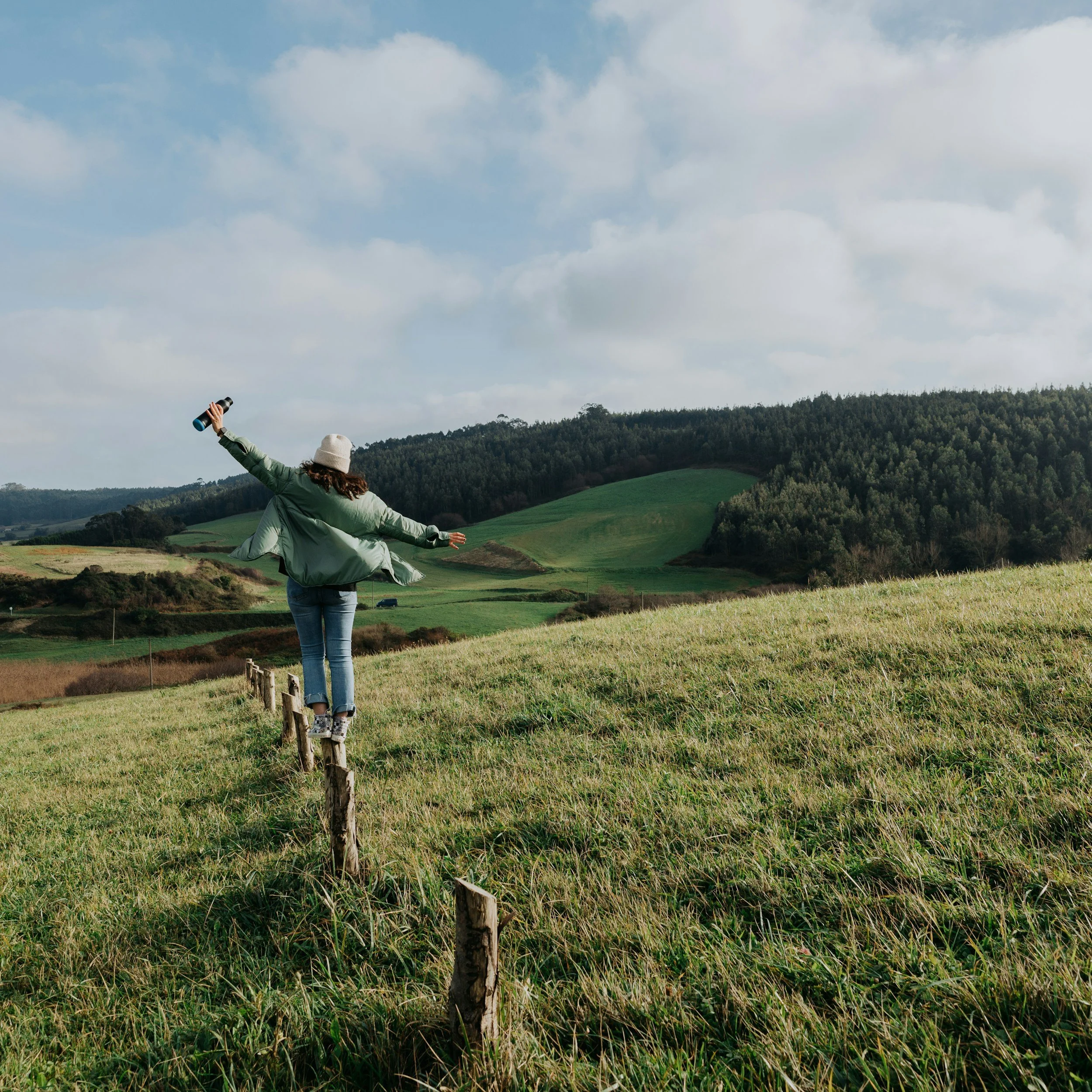 A person wearing a wide-brimmed hat and a green jacket is standing in a lush green field with their arms outstretched, holding binoculars in one hand, looking towards clouds over rolling hills and trees in the distance.