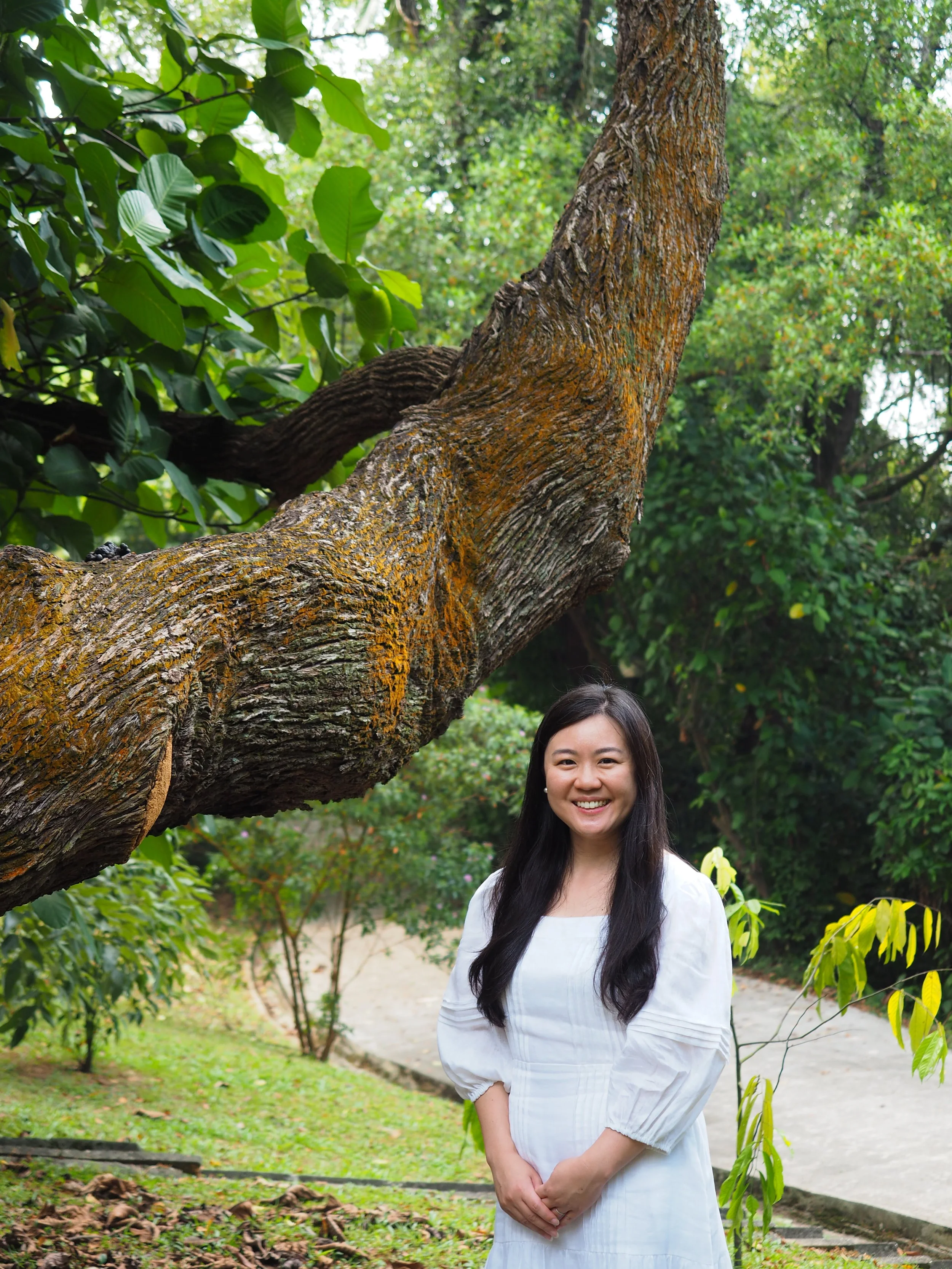 A young woman with long dark hair and a white dress standing outdoors in a lush garden. She is smiling and has her hands clasped in front of her, surrounded by green trees and plants with a path in the background.
