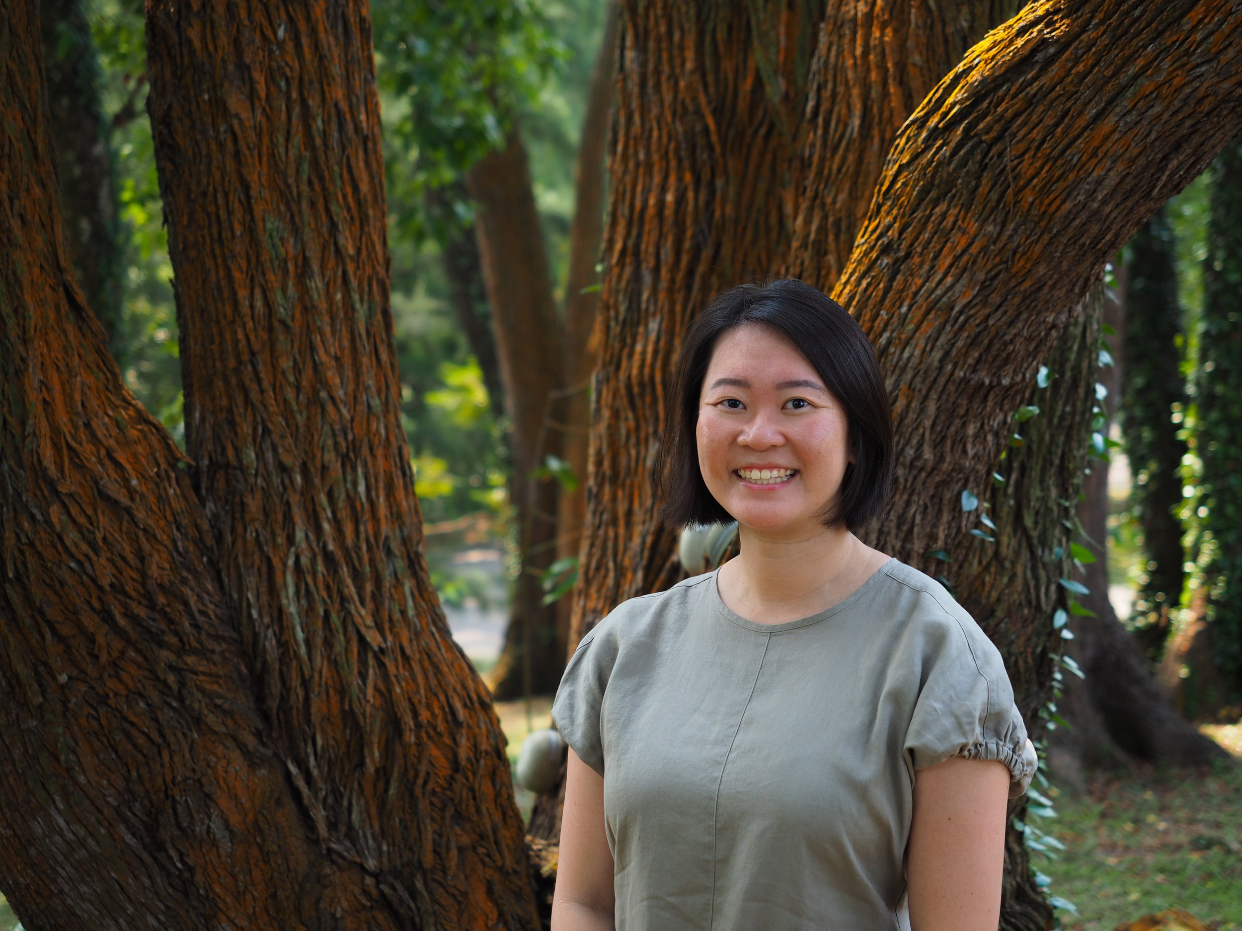 A woman with dark hair and a gray dress standing outdoors near large trees with rough bark, smiling at the camera.