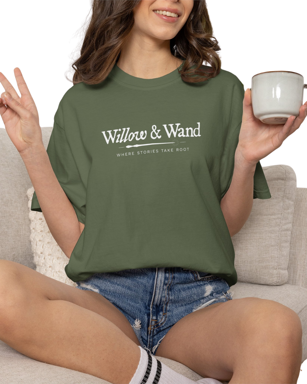 A woman sitting on a sofa, wearing a green t-shirt that says "Willow & Wand" with the tagline "Where Stories Take Root," and holding a white mug.