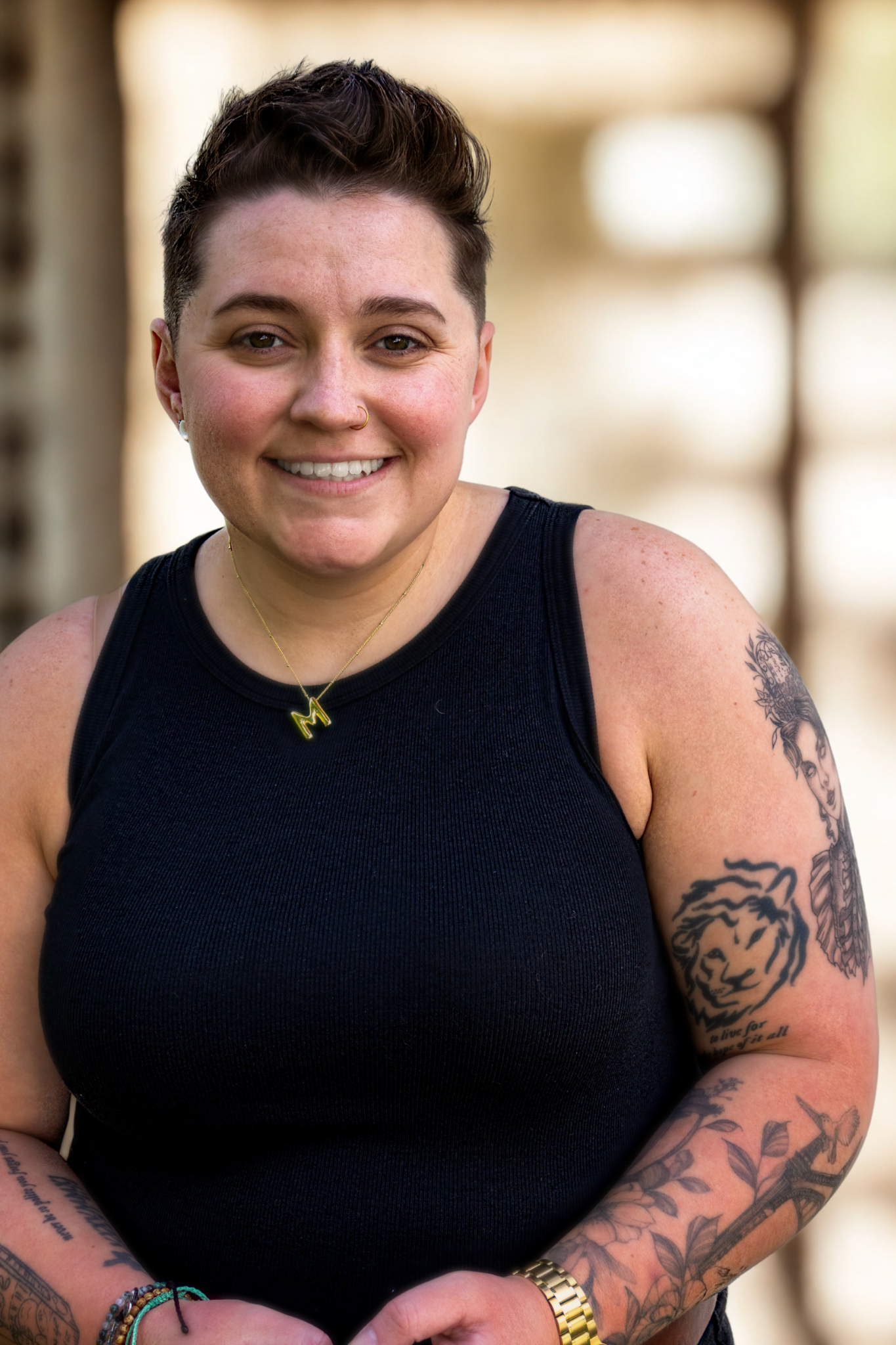 A young woman with short dark hair, wearing a black sleeveless top, gold necklace with a letter 'M', and bracelets on her left wrist. She has visible tattoos on her arms and has a friendly smile. The background is blurred and appears to be an outdoor setting with natural light.