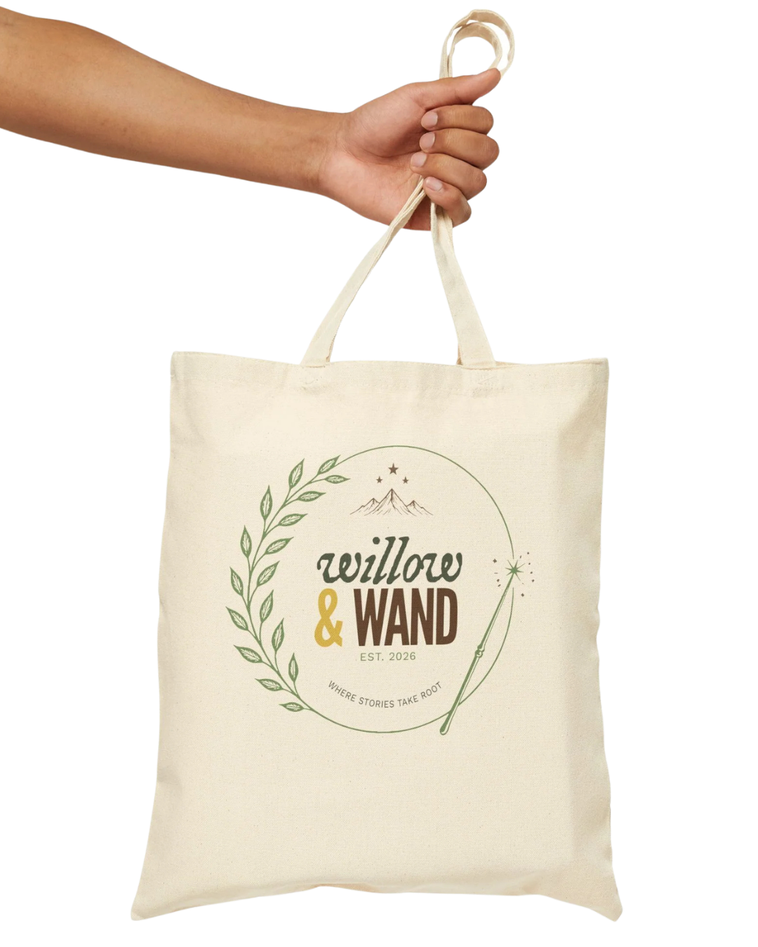 A hand holding a beige tote bag with the logo 'willow & wand' and the slogan 'where stories take root' printed on it.