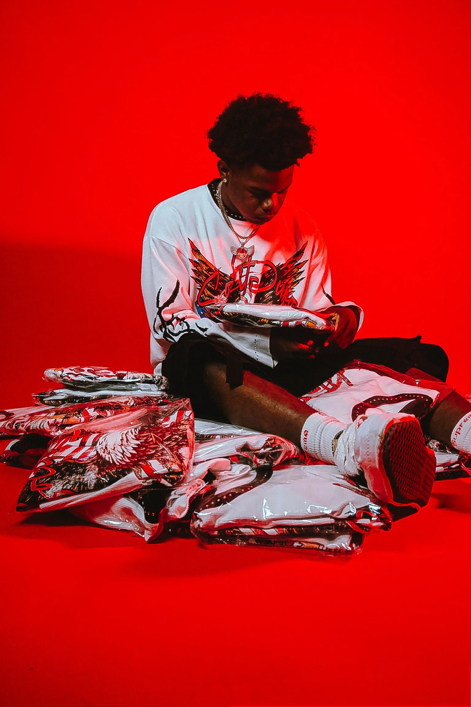 A young man with dark curly hair sitting cross-legged on a red background, surrounded by wrapped items with graphic designs.