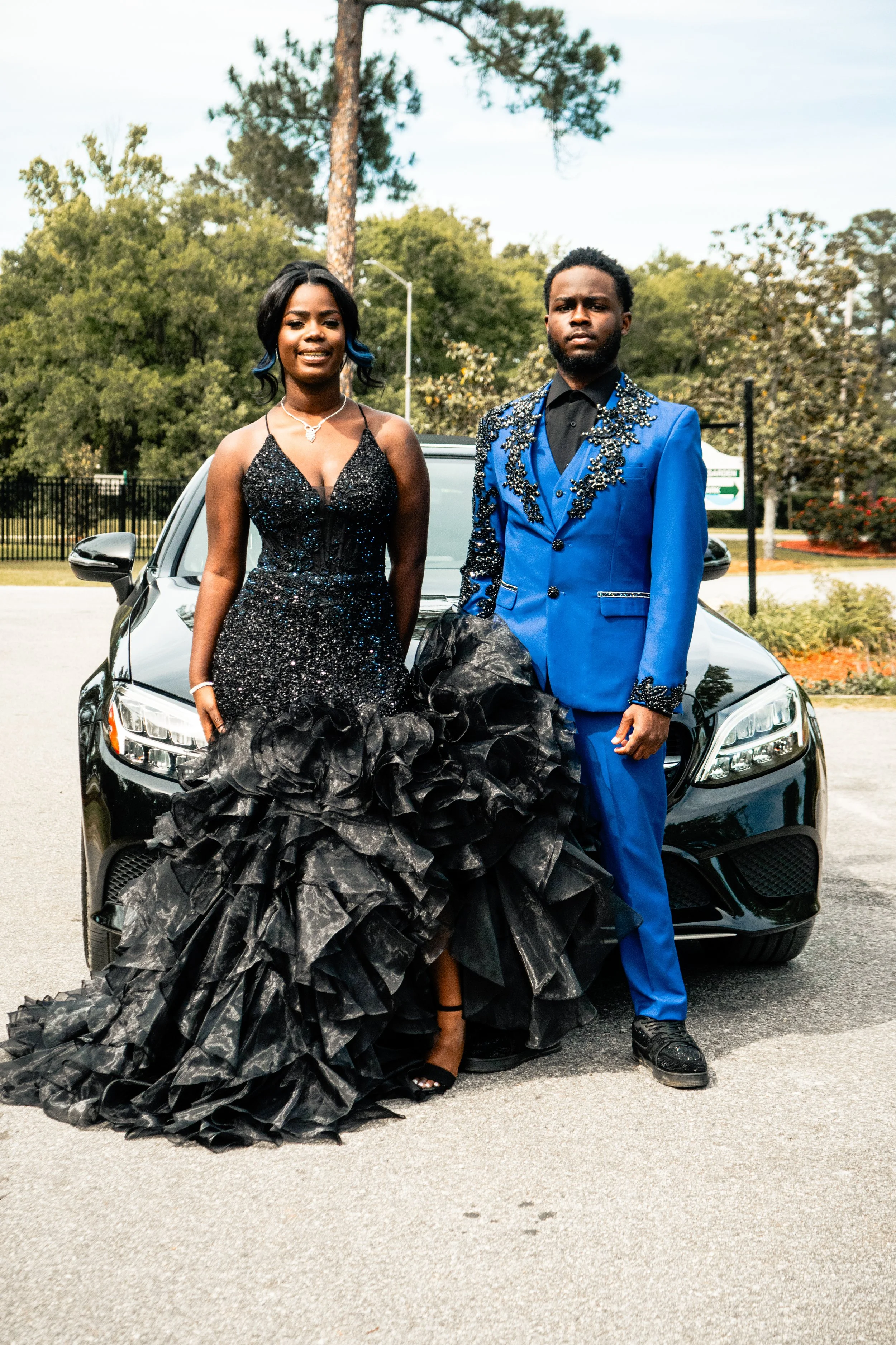 A couple dressed in formal attire standing in front of a black car outdoors. The woman is wearing a black, glittery gown with a ruffled skirt, and the man is in a bright blue suit with embellishments on the jacket. They are posing together with trees
