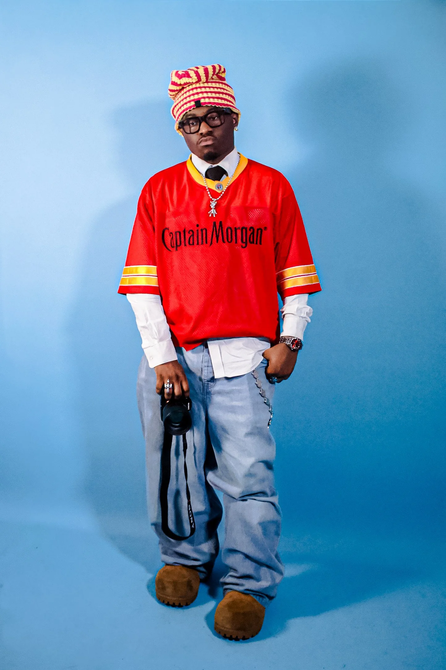 A young man wearing a red Captain Morgan shirt, layered over a white shirt, with a striped beanie, glasses, jewelry, and tan boots, holding a camera in his right hand against a blue background.