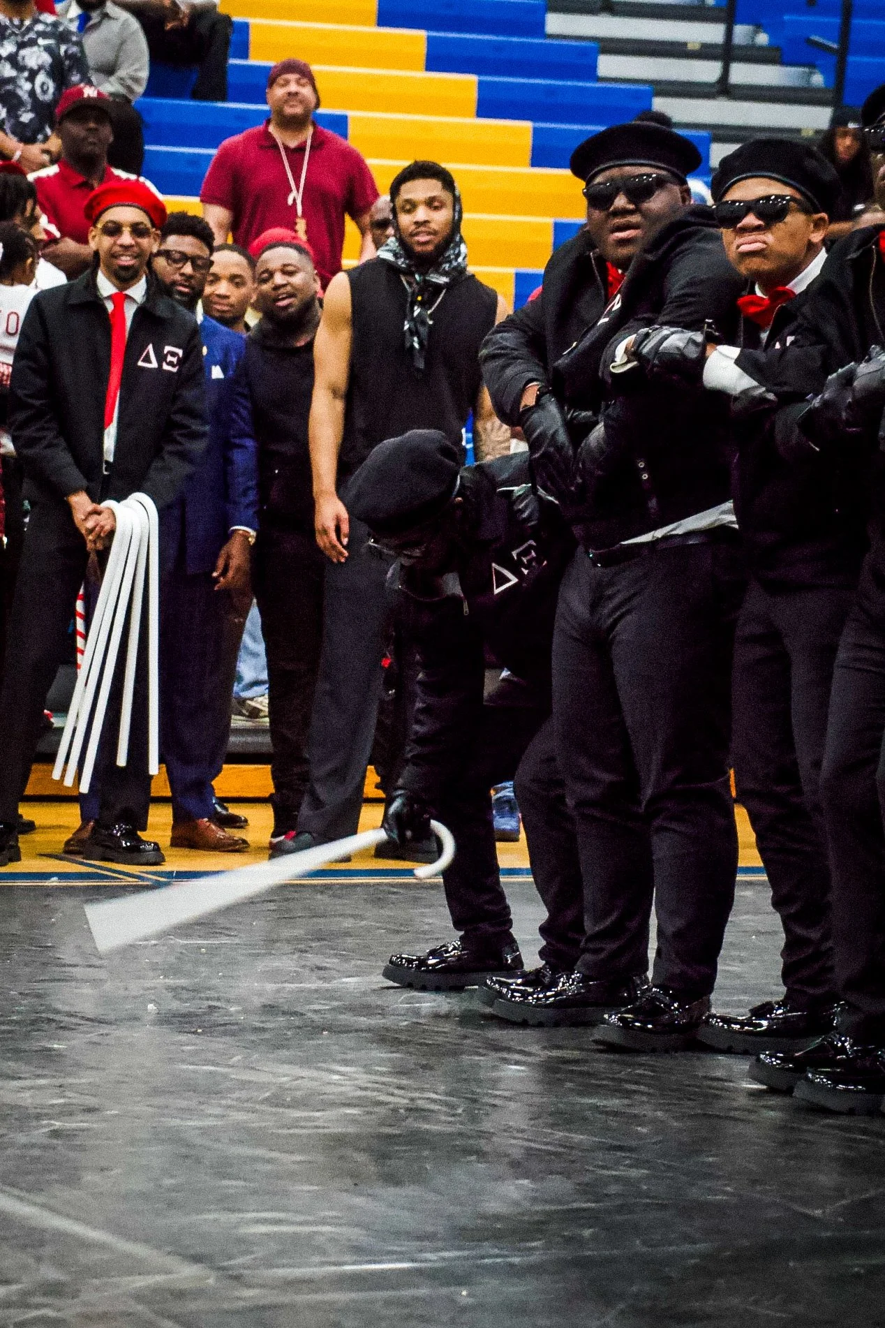 Group of men dressed in black suits, some with red ties, standing in a gymnasium with blue and yellow bleachers in the background. One man in the foreground is holding and swinging an axe, wearing reflective sunglasses and black gloves.