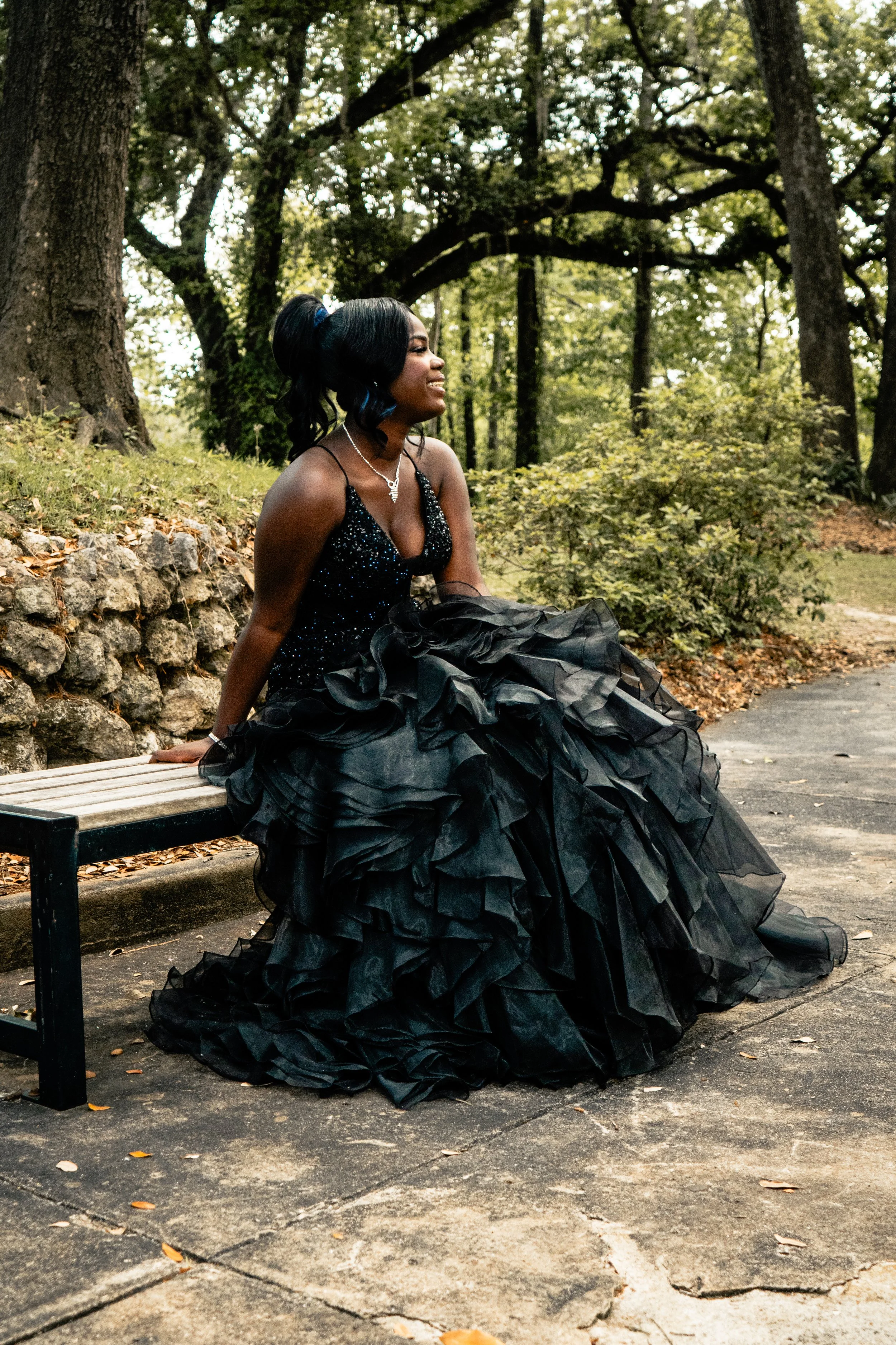 A woman in a black, ruffled ball gown sitting on a park bench, smiling and looking to her right, with tall trees and greenery in the background.