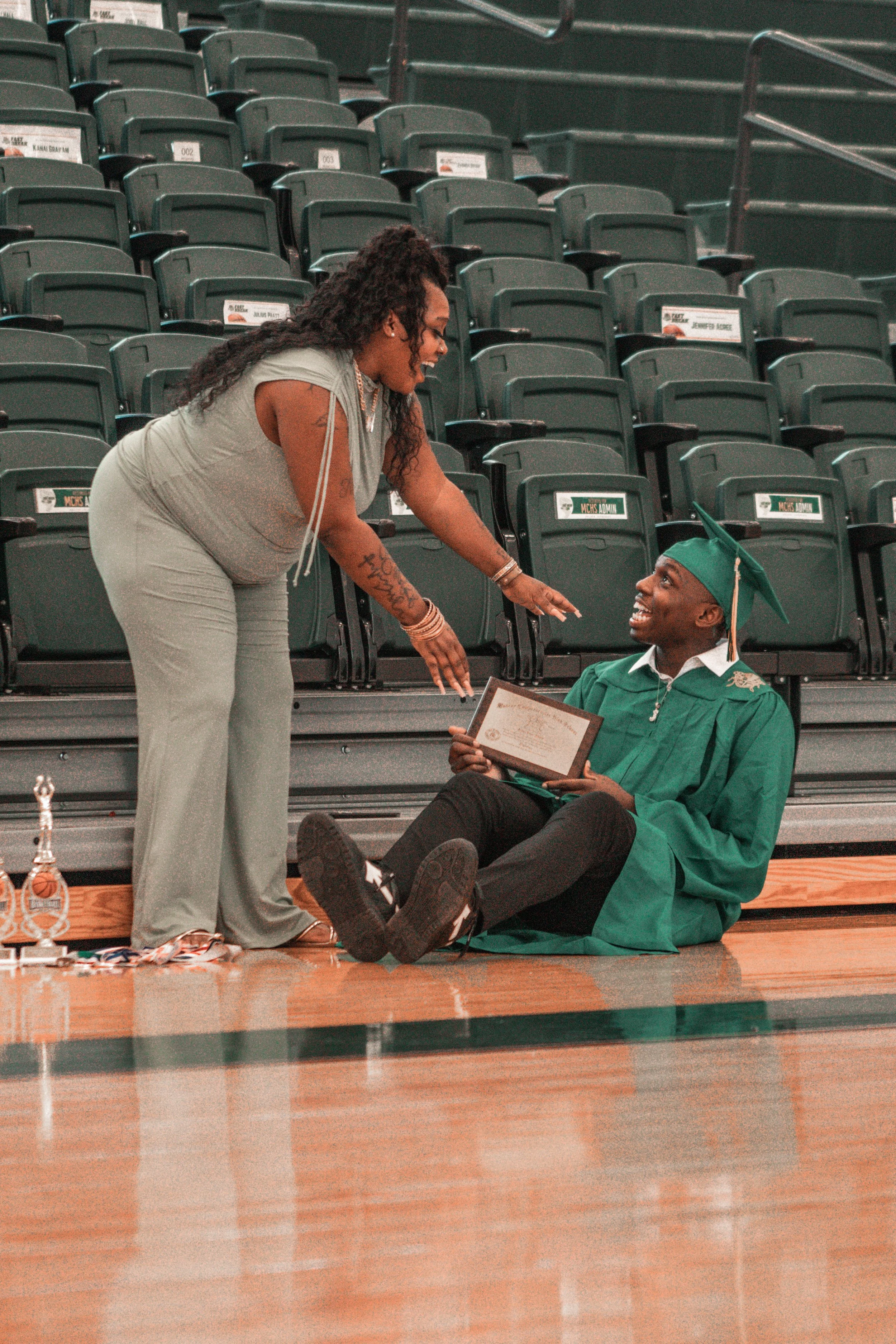 A woman leaning over a seated young man in graduation attire, both smiling, in an indoor arena with empty green seats.