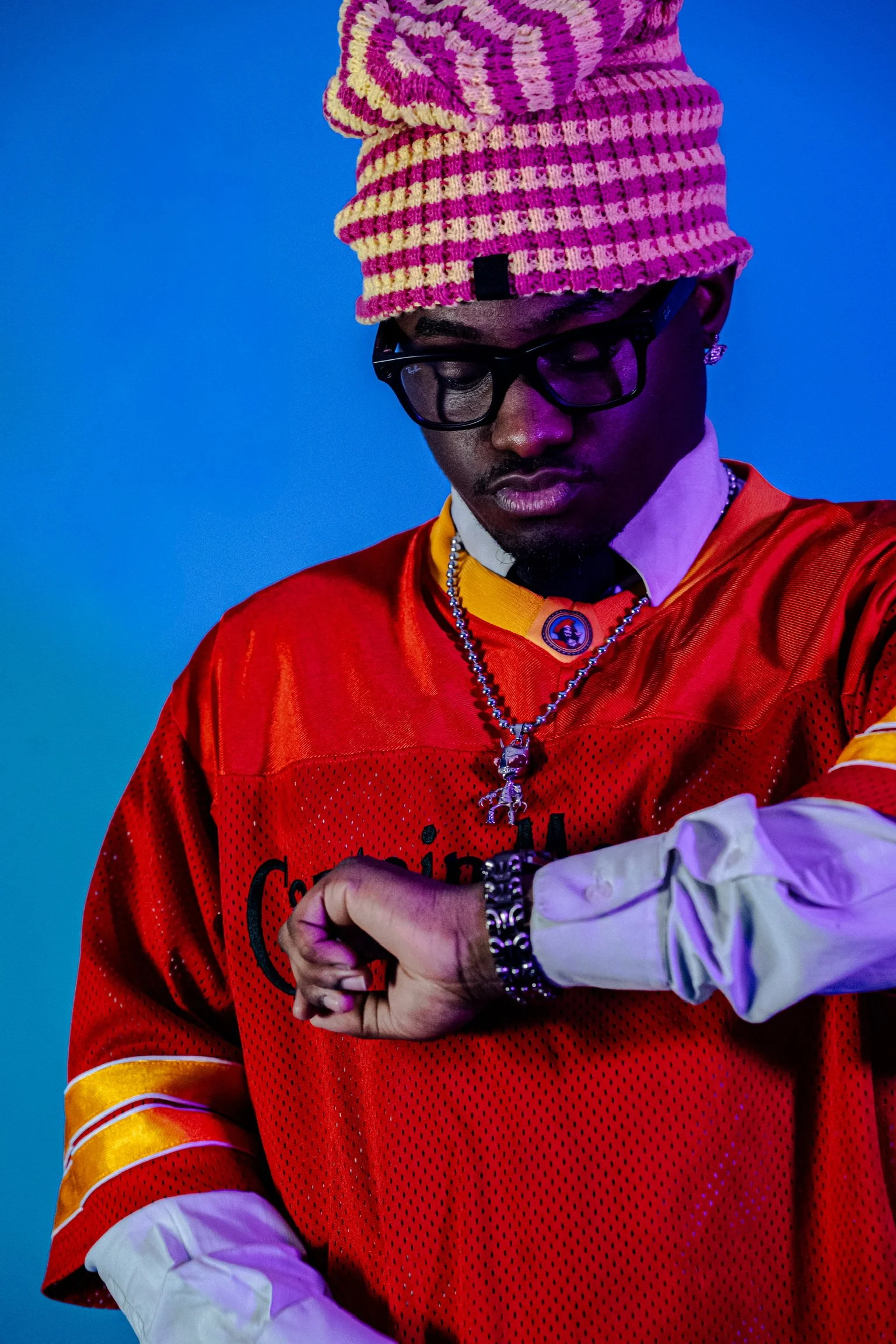 A man with dark skin wearing glasses, a multicolored beanie, and a red sports jersey. He has jewelry including a necklace, bracelet, and earrings. The background is blue.