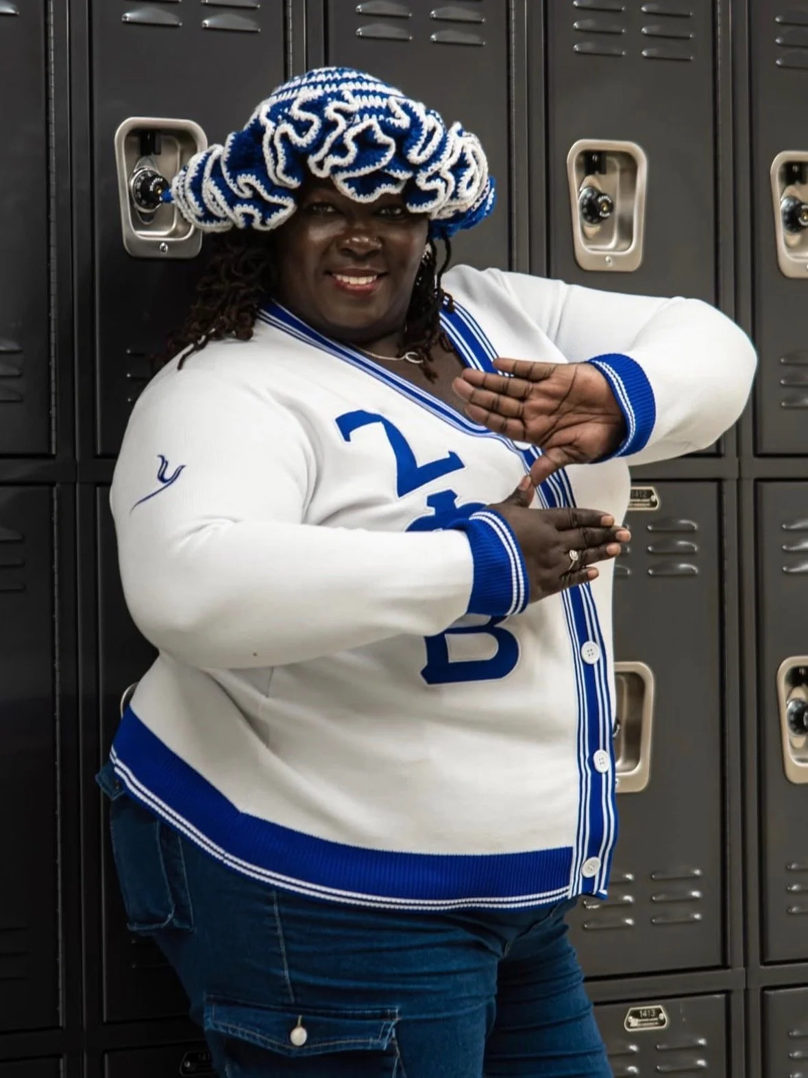 A woman standing in front of lockers wearing a white and blue letterman jacket, a knitted hat with blue and white stripes, and making a gesture with her hands.